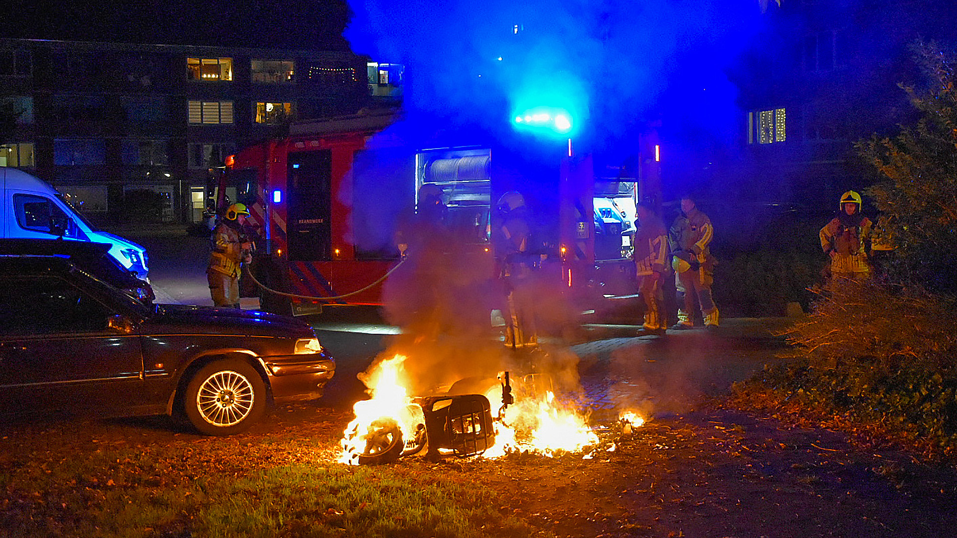 Brandweerlieden blussen een brandende elektrische scooter bij nacht, met een brandweerwagen op de achtergrond.