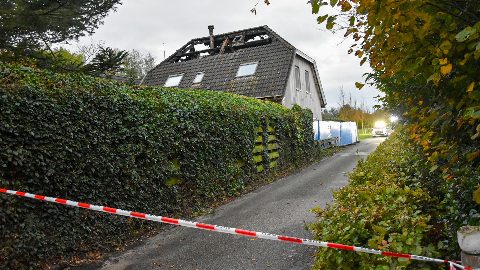 Een huis met een beschadigd dak, omringd door een heg, met politieafzetting ervoor en een op de achtergrond zichtbare politieauto met zwaailichten.