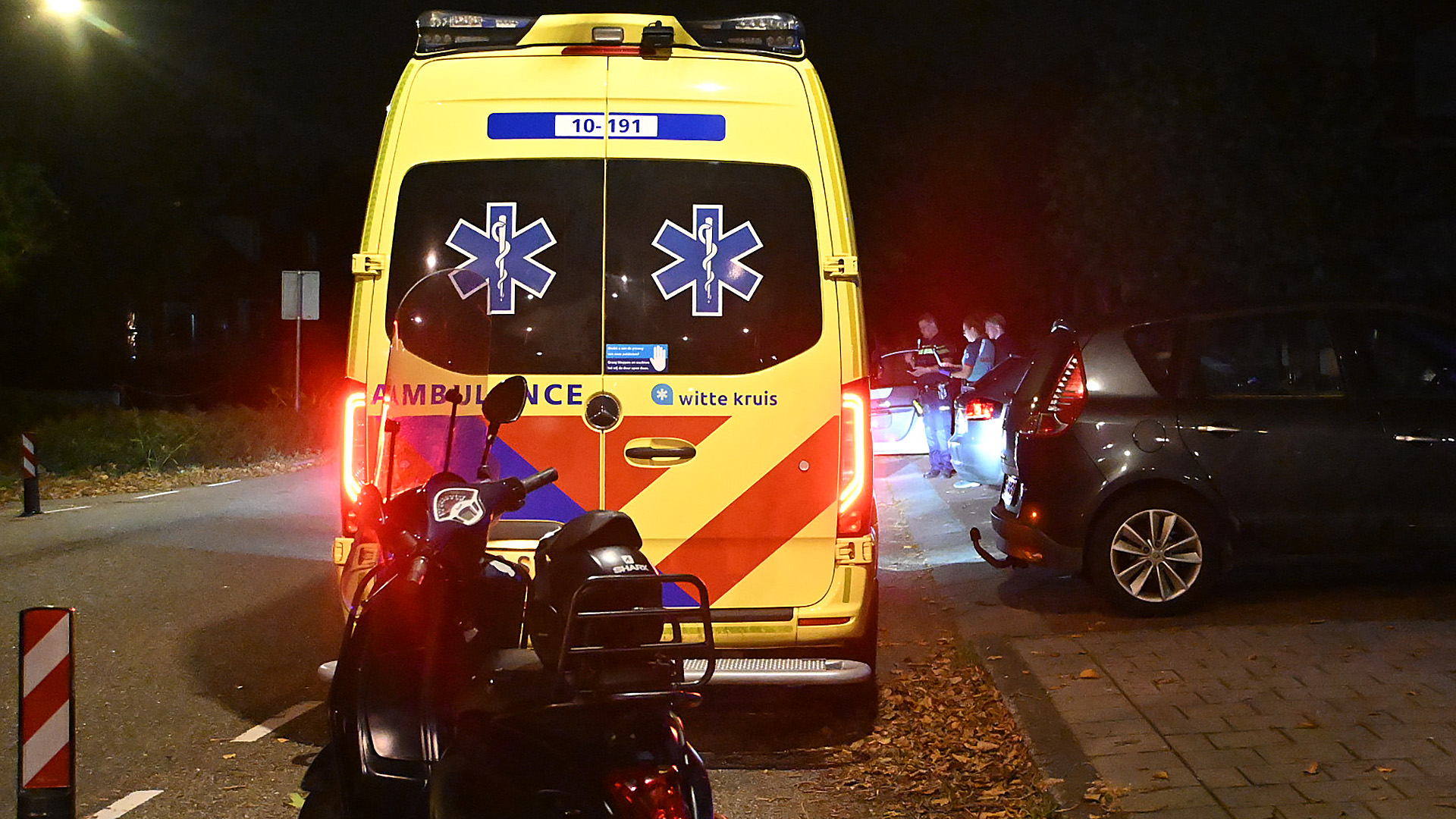Een ambulance staat 's nachts op een weg, met politieagenten bij een auto verderop en een scootmobiel op de voorgrond.