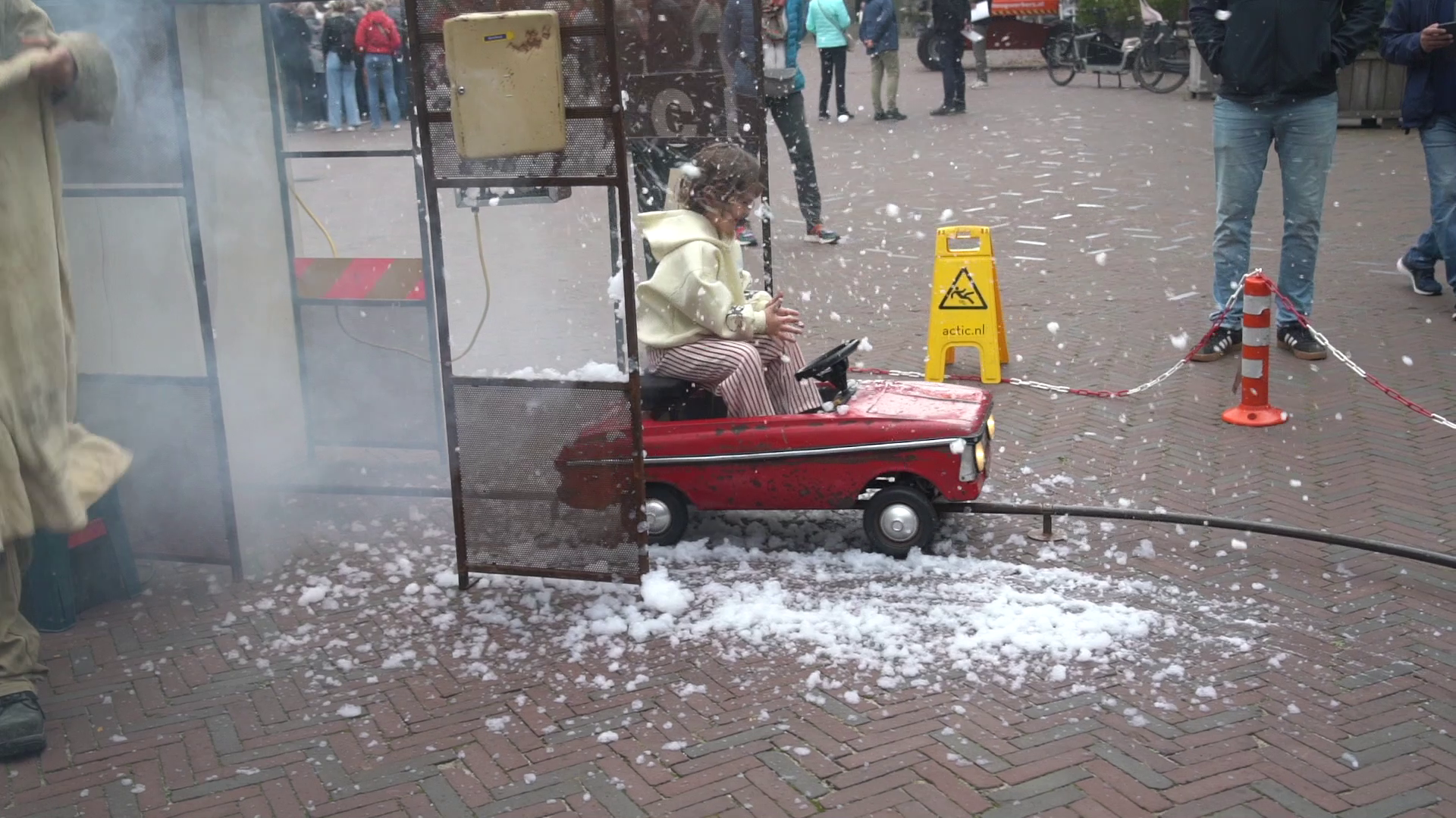 Een jong kind in een rode trapauto tijdens een straatvoorstelling, omringd door schuim en rook. Op de achtergrond staan enkele toeschouwers en een waarschuwingsbord voor gladde vloer.