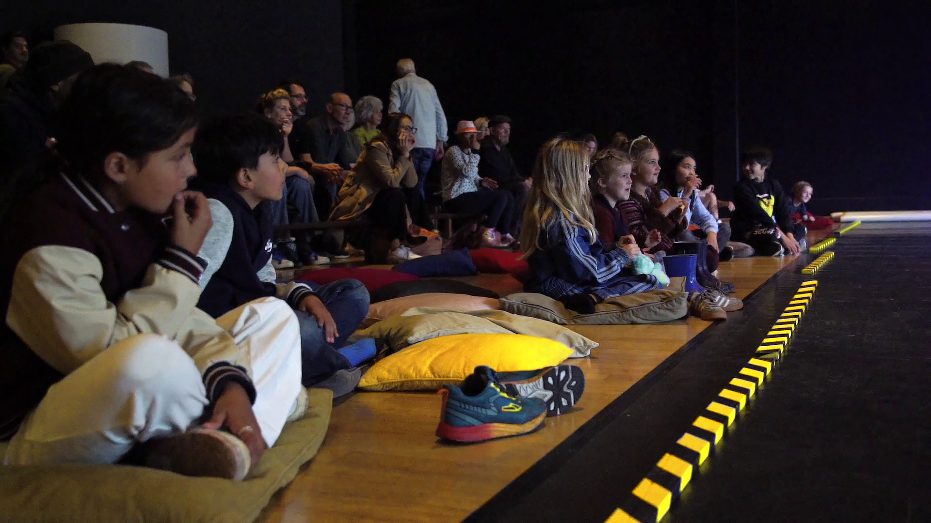 Kinderen zitten op kussens op de vloer in een zaal, aandachtig kijkend naar een optreden of voorstelling.
