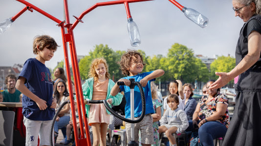 Kinderen spelen met een interactieve installatie met plastic flessen onder begeleiding van een volwassene, omringd door toeschouwers in een buitenomgeving op een zonnige dag.