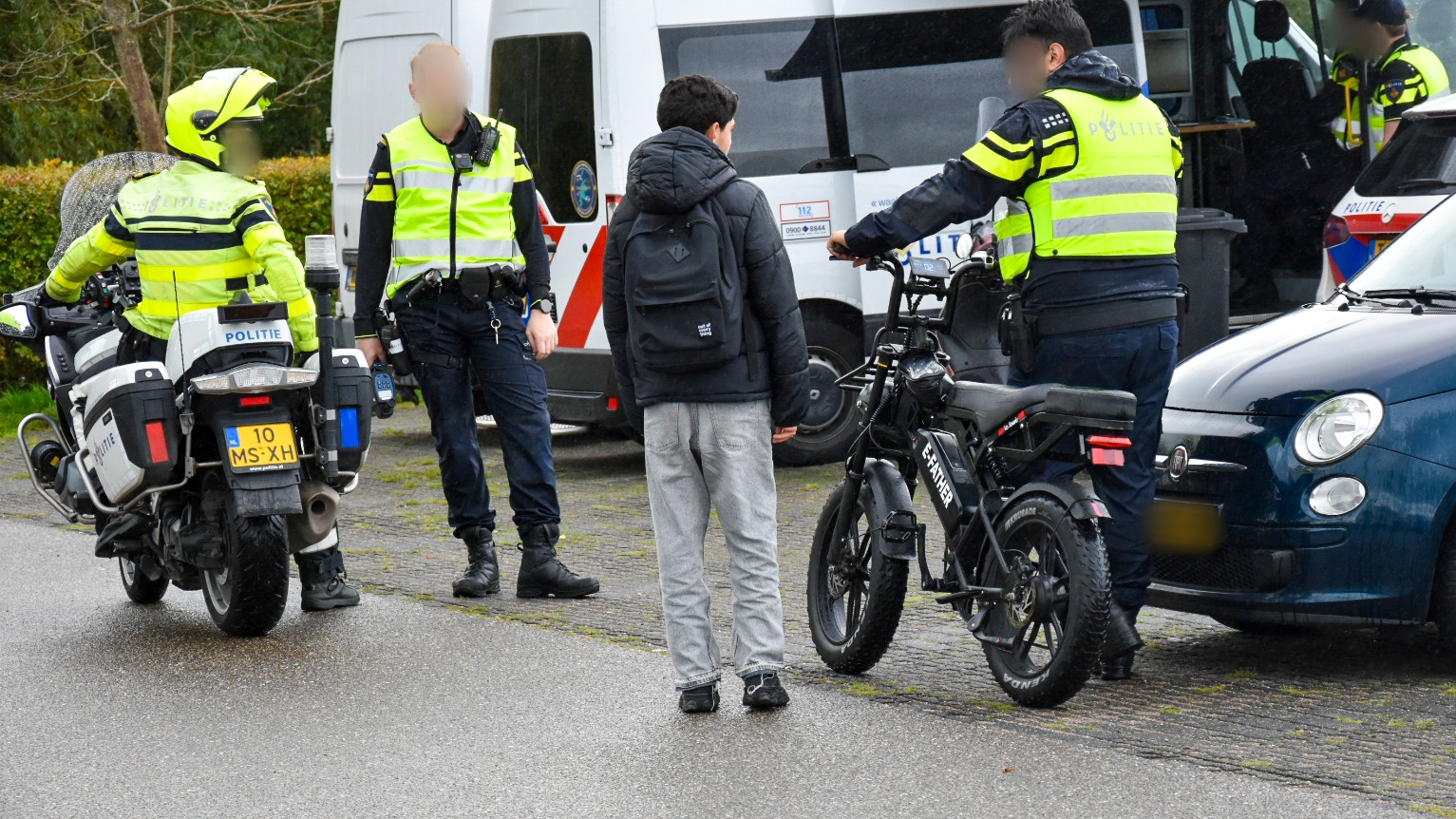 Twee politieagenten in reflecterende jassen praten met een jongeman op een parkeerplaats naast een elektrische fiets en een motorfiets. Een politieauto staat geparkeerd op de achtergrond.