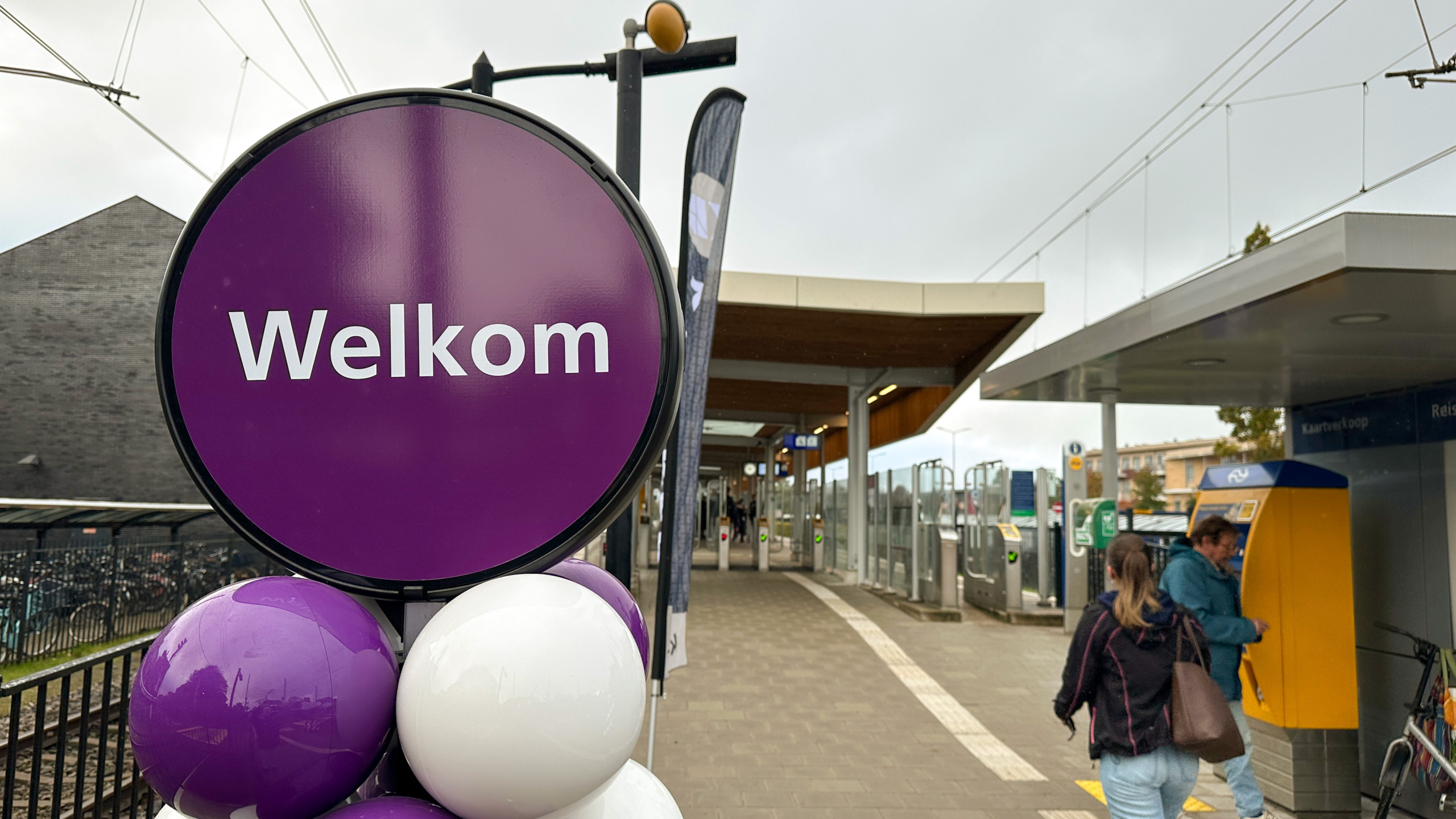 Een paarse welkomstbord staat voor een treinstation met ballonnen en mensen bij een kaartautomaat.
