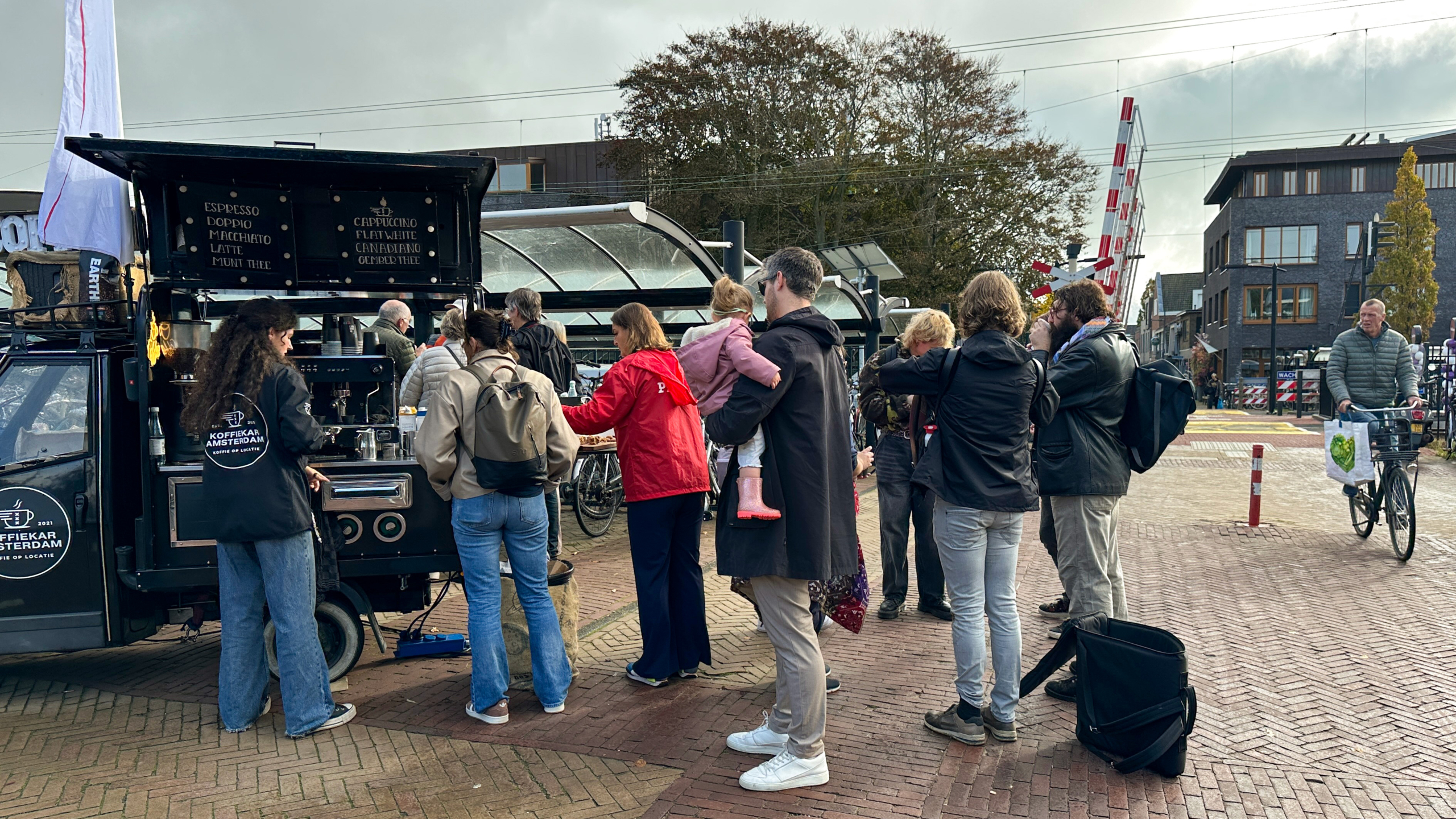 Mensen staan in de rij voor een mobiele koffiekar in een stedelijke omgeving, met fietsenstalling en spoorweg overweg op de achtergrond.