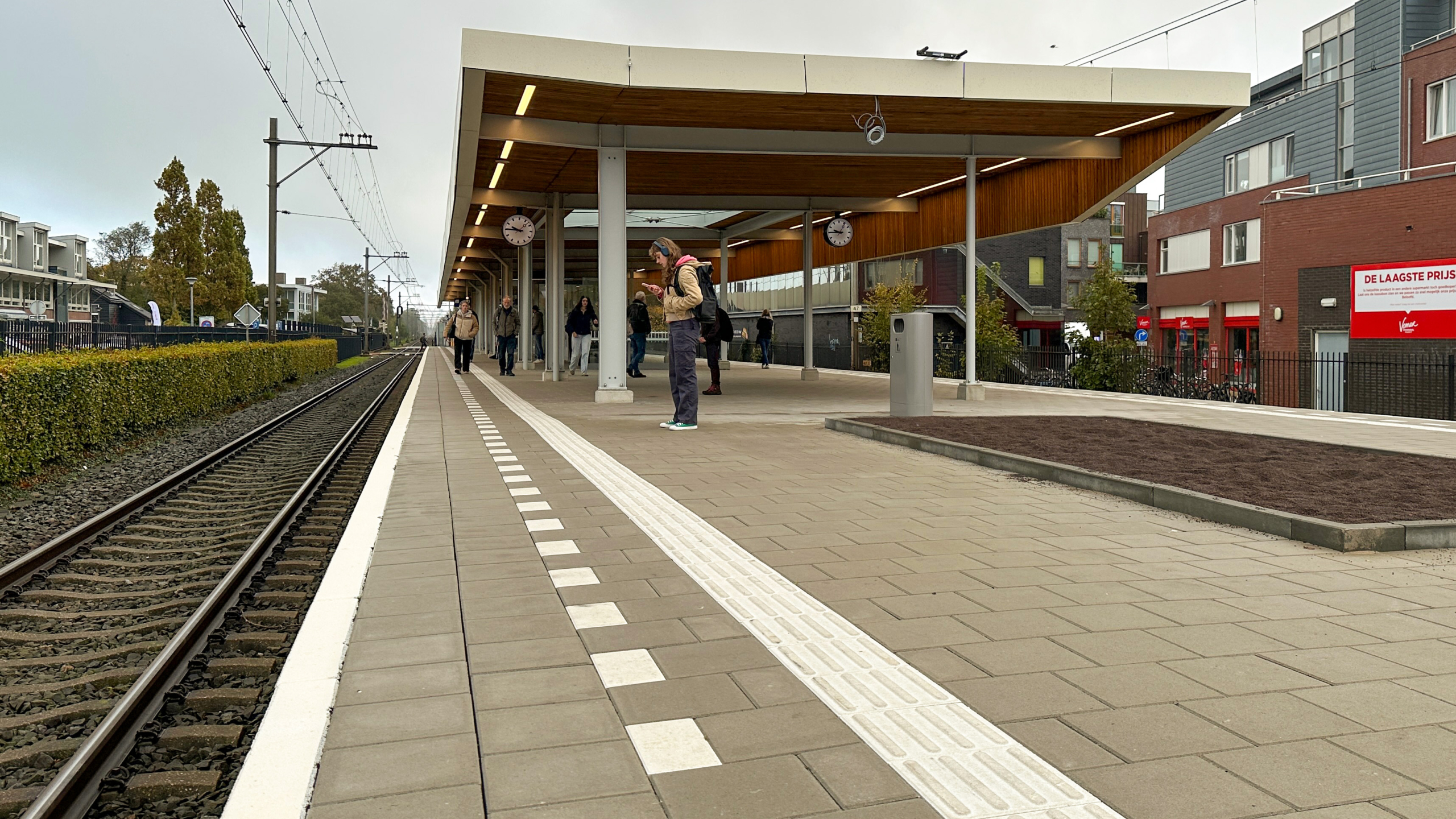 Een treinperron met mensen die rondlopen of wachten, met een overkapping en stationsklokken aan het plafond. Links van het perron zijn treinsporen te zien. Op de achtergrond staan moderne gebouwen en een hek met fietsen.