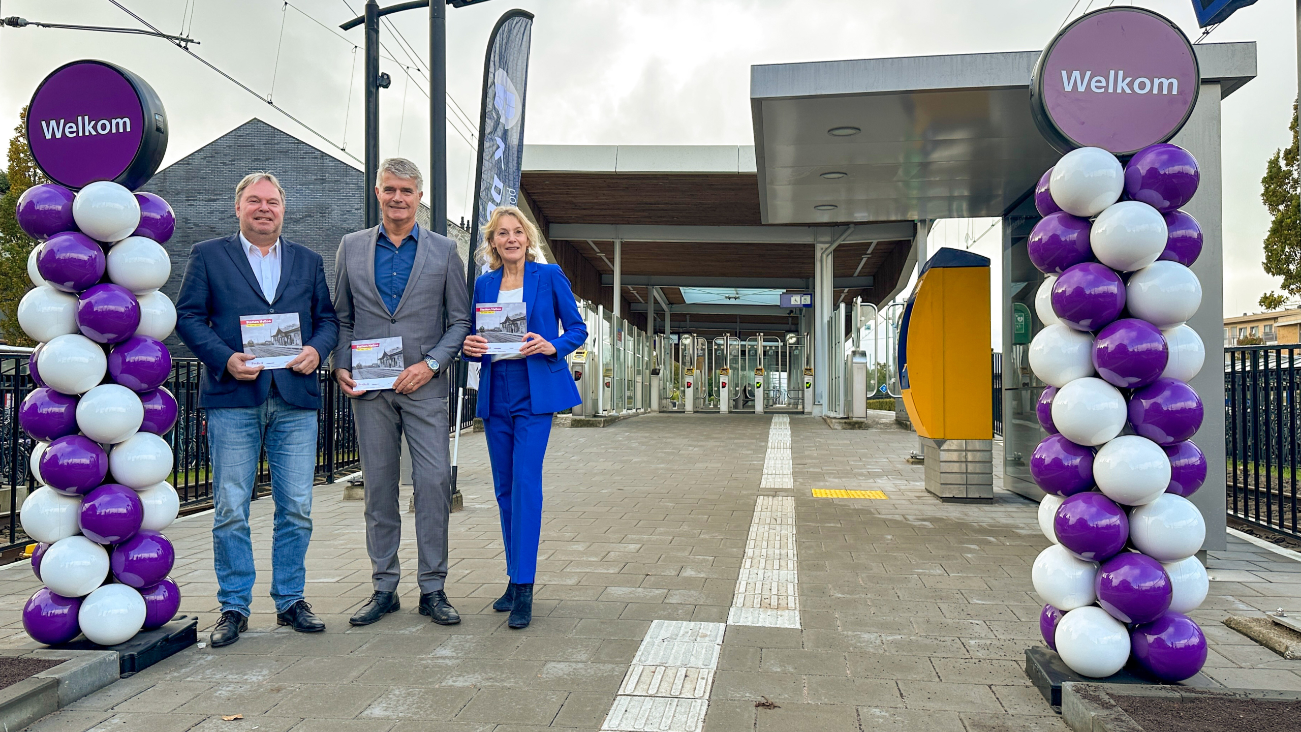 Drie mensen poseren voor een stationingang versierd met paarse en witte ballonnen en borden met "Welkom" erop.