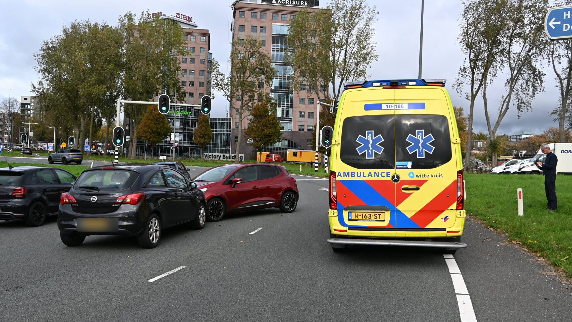 Een ambulance staat aan de kant van de weg, terwijl meerdere auto's in een lichte botsing betrokken lijken te zijn. Op de achtergrond zijn hoge kantoorgebouwen zichtbaar en een persoon staat op het gras met een mobiele telefoon.
