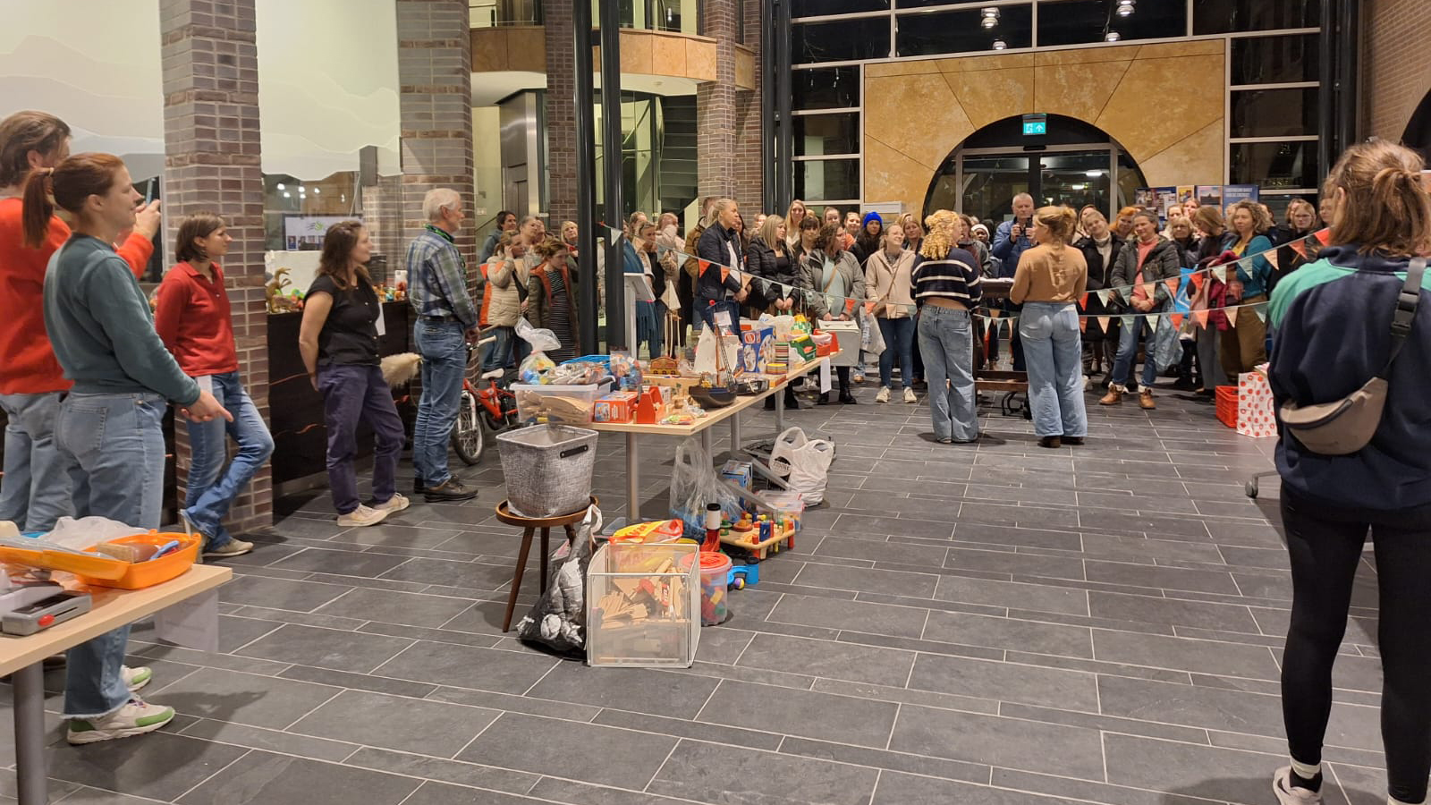 Een grote groep mensen verzamelt zich binnen in een ruime hal, waar tafels vol speelgoed en andere spullen staan uitgestald. Er hangt een kleurrijke slinger boven de tafels, en de sfeer lijkt druk maar gemoedelijk.