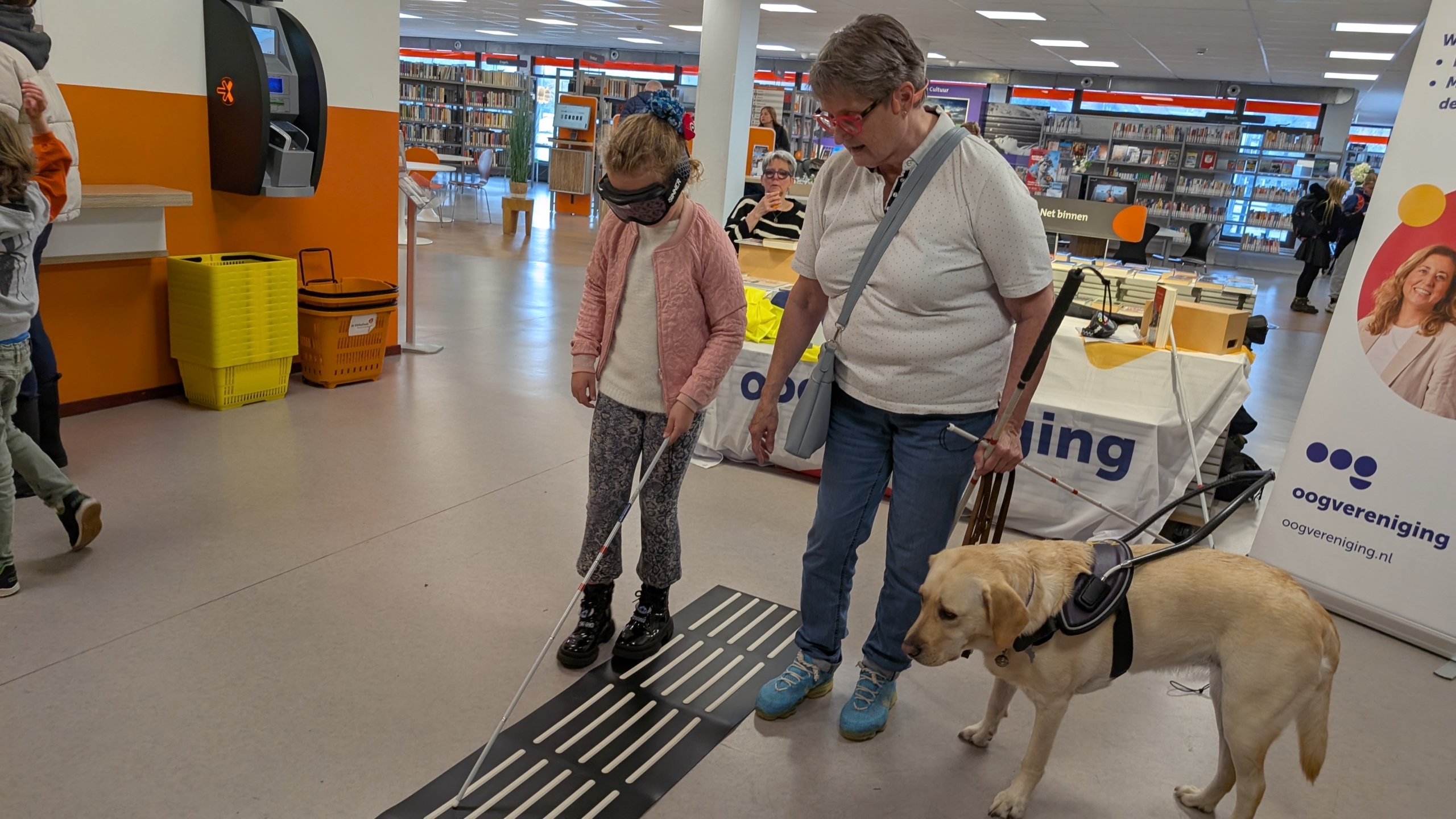 Een jong meisje met een blinddoek en taststok loopt op een geautomatiseerde gidsbaan in een bibliotheek, begeleid door een oudere vrouw met een hondengeleidehond.