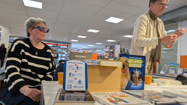 Een vrouw met een donkere zonnebril en gestreepte trui zit aan een tafel in een bibliotheek, met naast haar een man die iets vasthoudt. Op de tafel liggen verschillende boeken, waaronder eentje met de titel 'Geleidehonden'. Boven de boeken is een bordje met informatie over blindenstokken zichtbaar.
