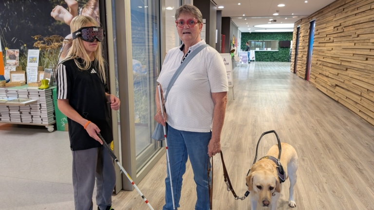 Een jonge vrouw met speciale bril en een oudere vrouw met witte stok en geleidehond staan in een winkelcentrum.