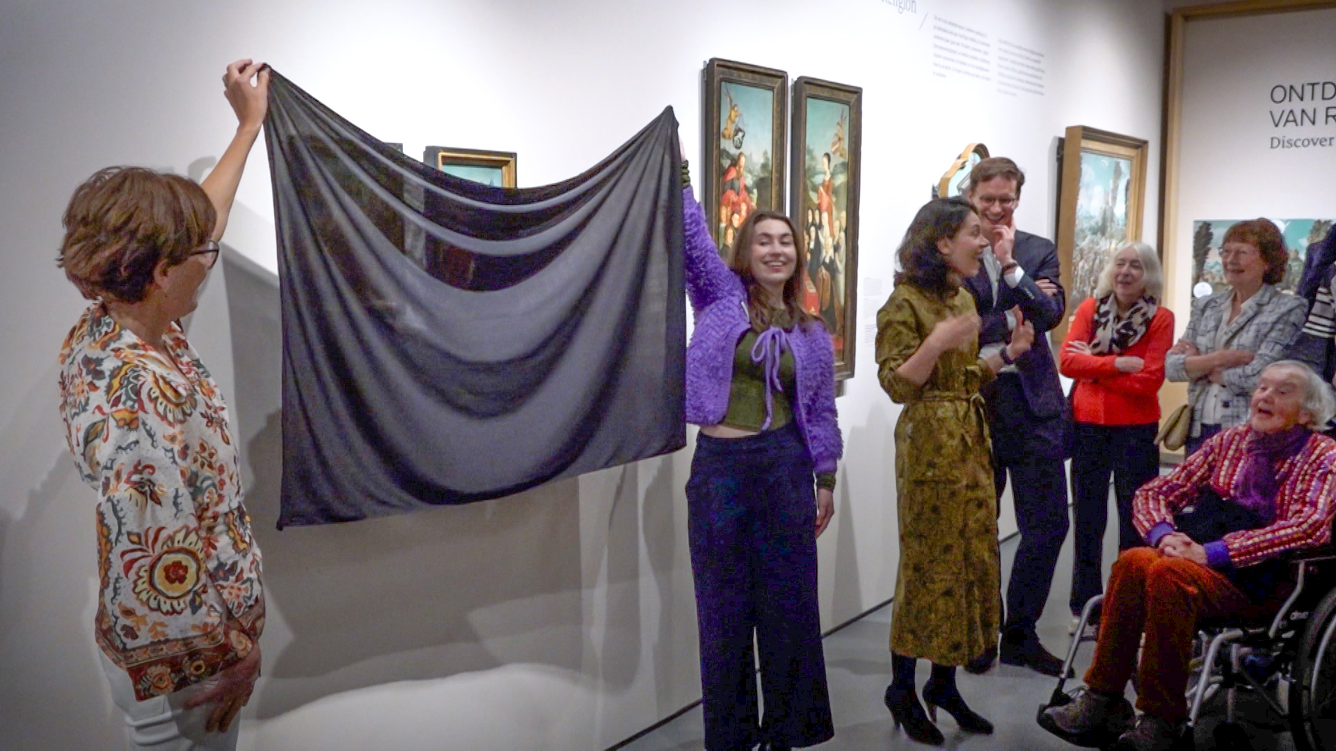 Een groep mensen verzameld in een museum terwijl een zwart doek gedeeltelijk wordt opgetild om een kunstwerk te onthullen. Er zijn schilderijen aan de muur te zien en een man in een rolstoel aan de rechterkant van de afbeelding.