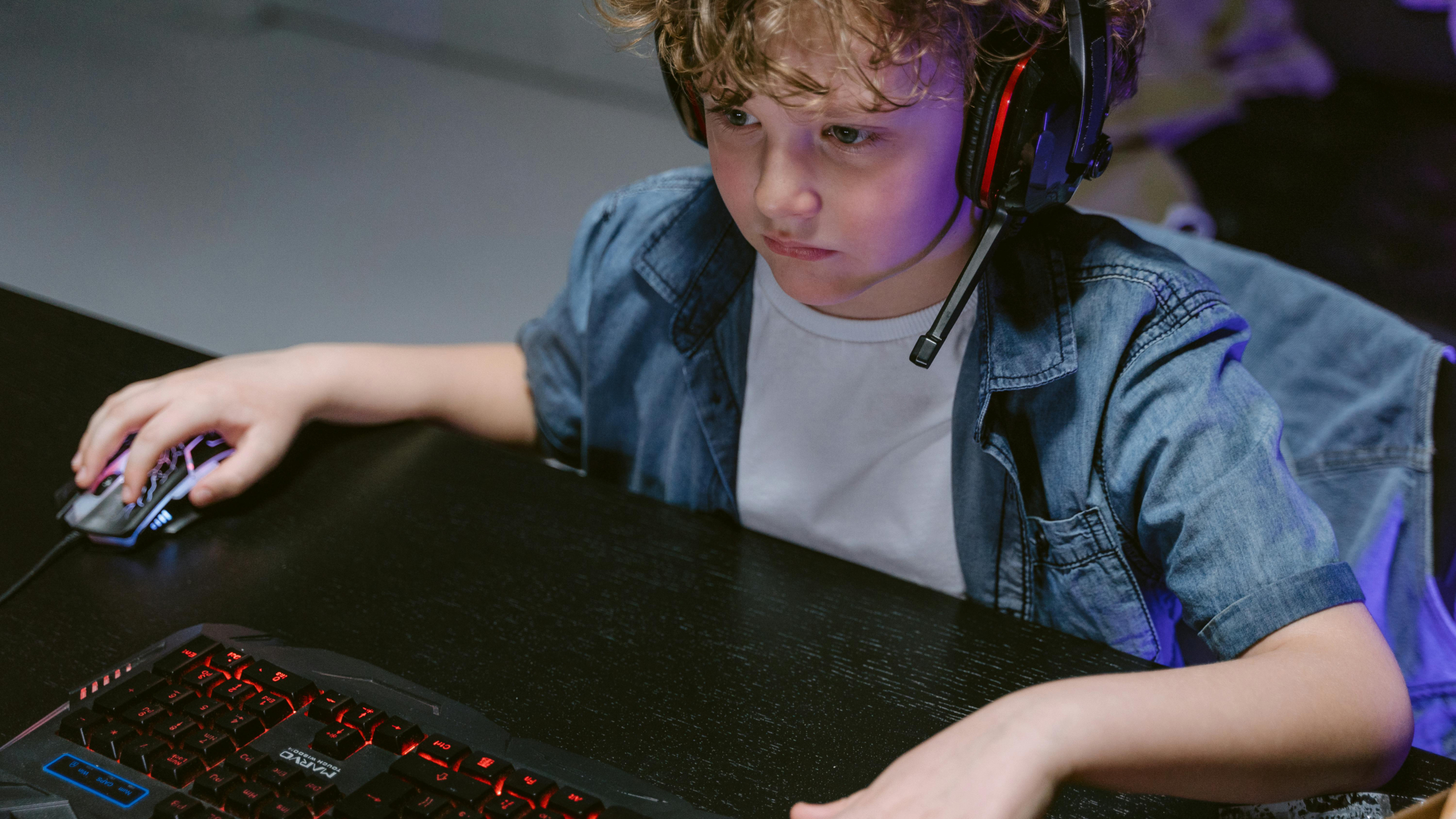 Jong kind met een koptelefoon zit geconcentreerd voor een computer en gebruikt een muis en keyboard met rode verlichting.