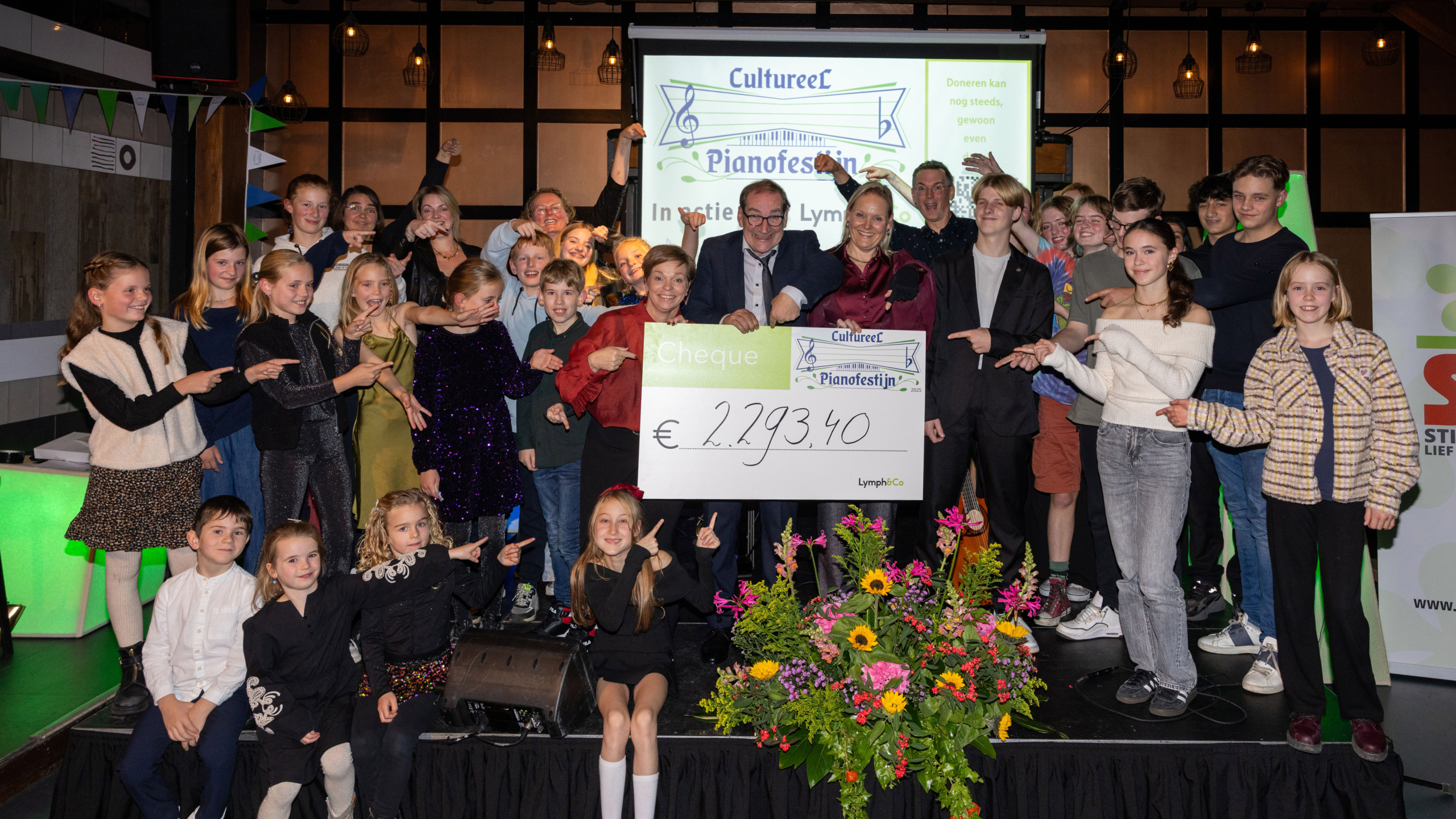 Een groep mensen houdt een grote cheque vast met het bedrag van €2.293,40 erop, omringd door bloemen. Er hangt een spandoek van 'Cultureel Pianofestijn' op de achtergrond.