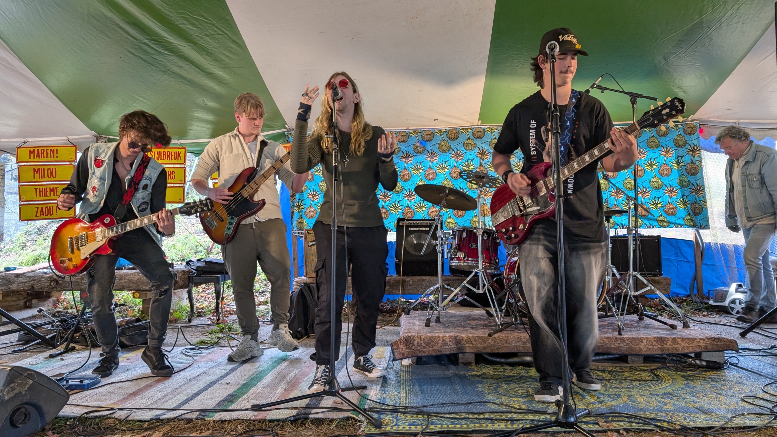 Een band speelt muziek op een podium onder een tent, met drie gitaristen en een drummer, waarbij de zanger in het midden in de microfoon zingt. De achtergrond is kleurrijk gedecoreerd met een patroon.