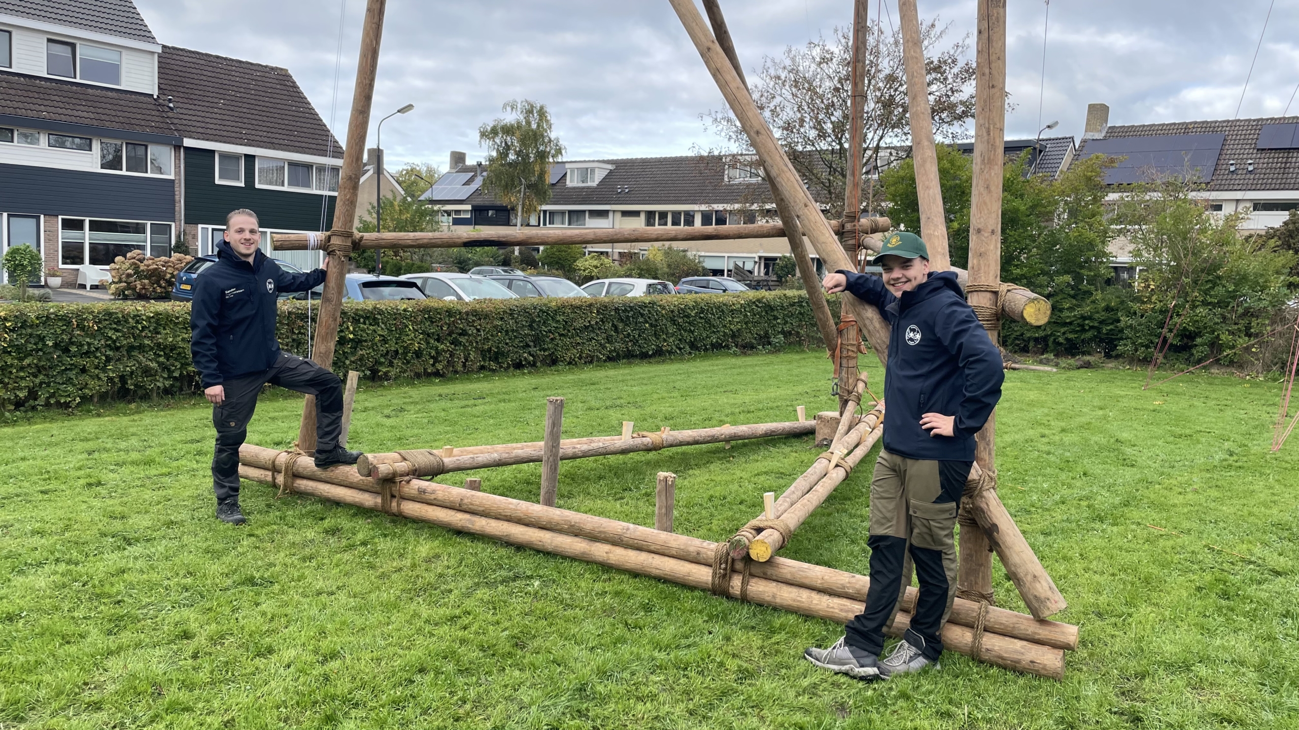 Twee mensen in scoutingkleding poseren naast een grote houten constructie op een grasveld, met huizen en auto's op de achtergrond.