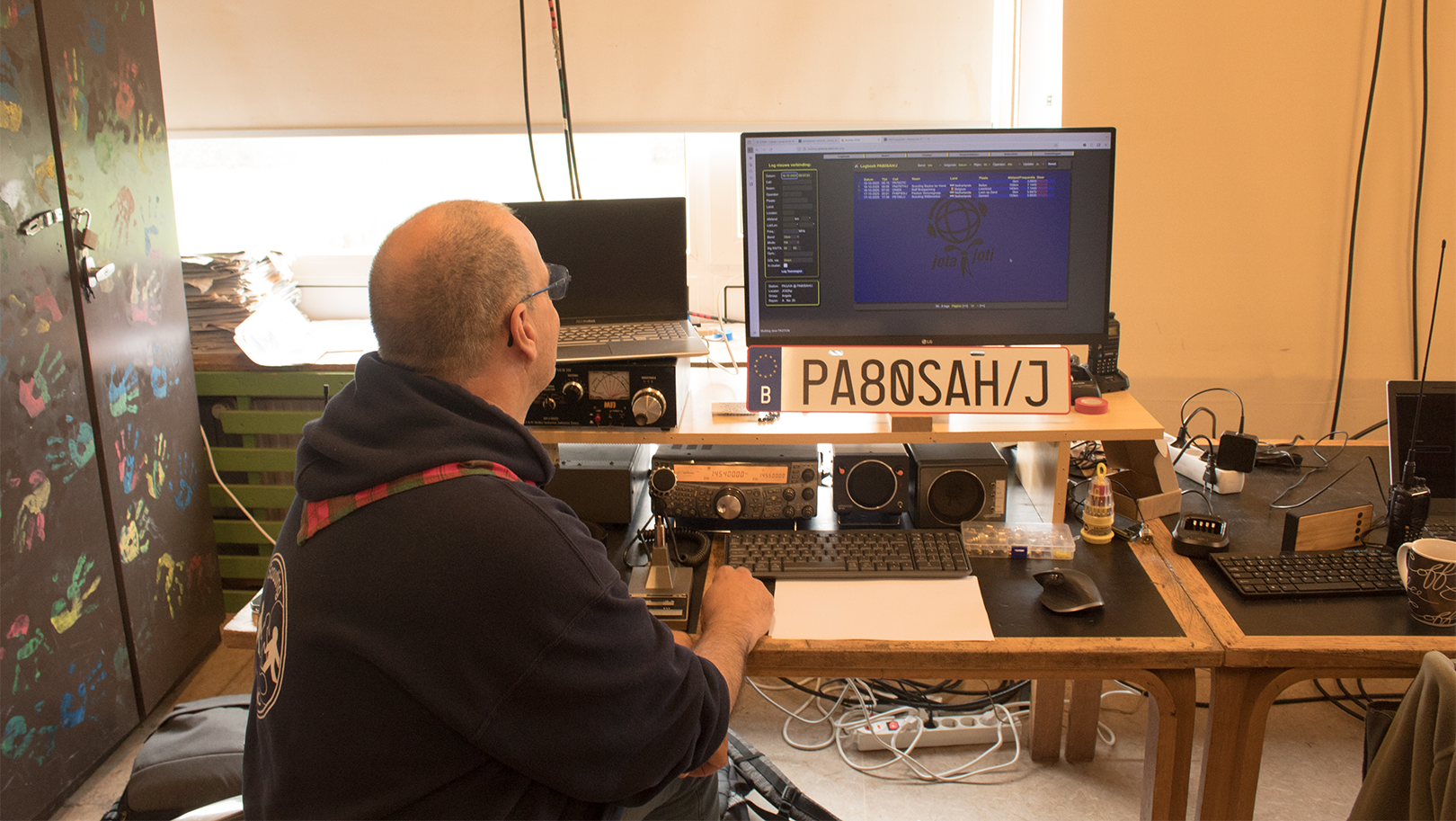 Een man in een donkere hoodie kijkt naar een computerscherm met radiocommunicatieapparatuur op een bureau. De ruimte heeft een kleurrijk beschilderde kast en een bord met het opschrift 'PA8ØSAH/J' staat prominent op het bureau.