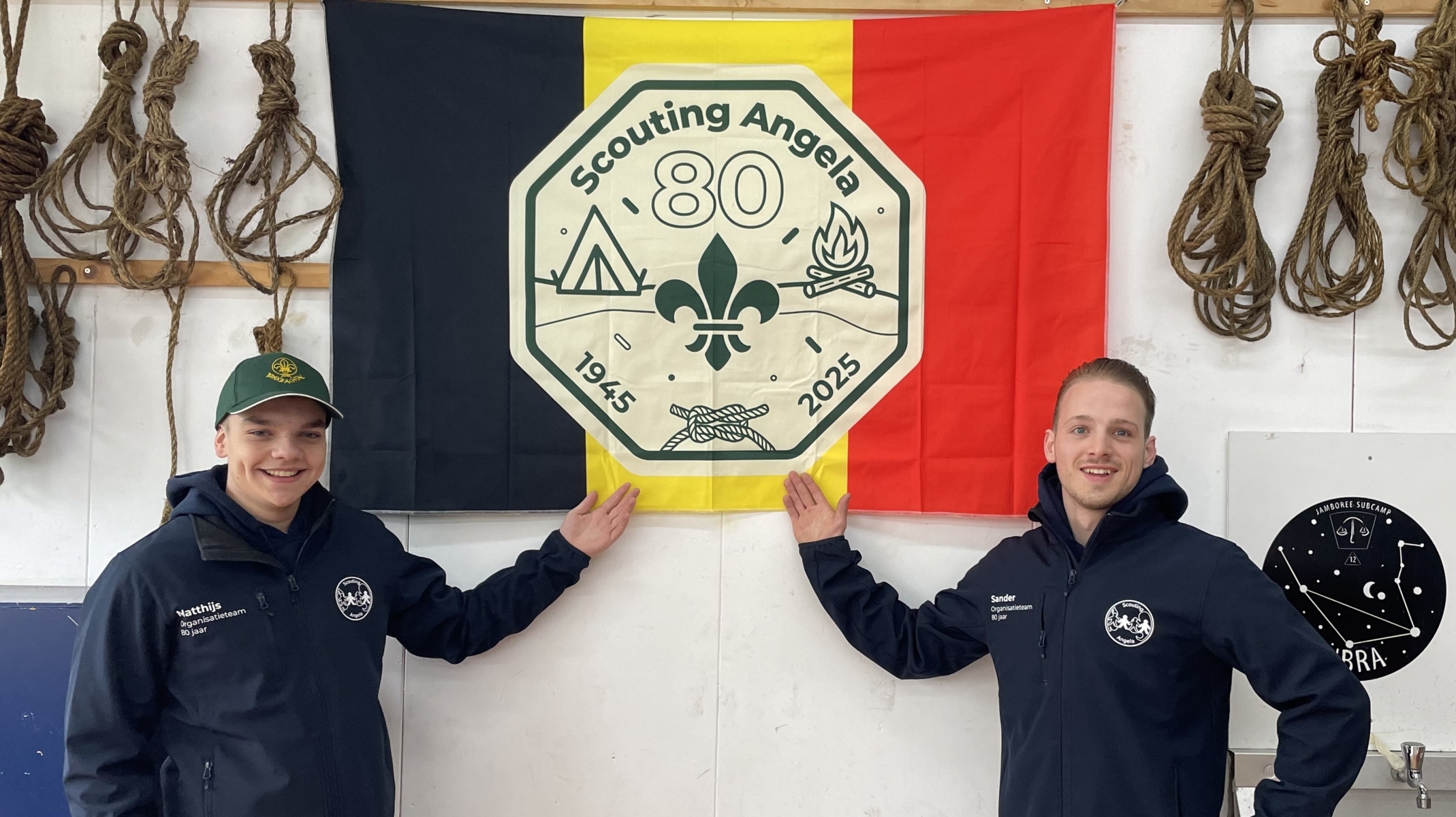 Twee mannen in scoutingkleding staan voor een Belgische vlag met het embleem van Scouting Angela, ter gelegenheid van het 80-jarig jubileum. Links hangen meerdere geknoopte touwen aan de muur.