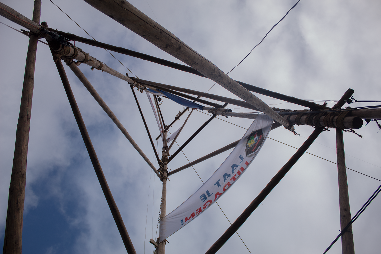 Onderkant van een hoge houten structuur met touwen en een wapperende vlag tegen een bewolkte hemel.