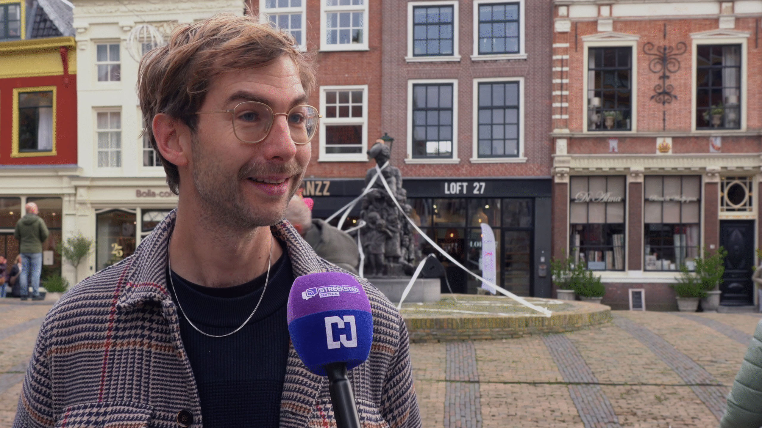 Man met bril wordt geïnterviewd, met microfoon van Streekstad Centraal, op een plein met historische gebouwen op de achtergrond.