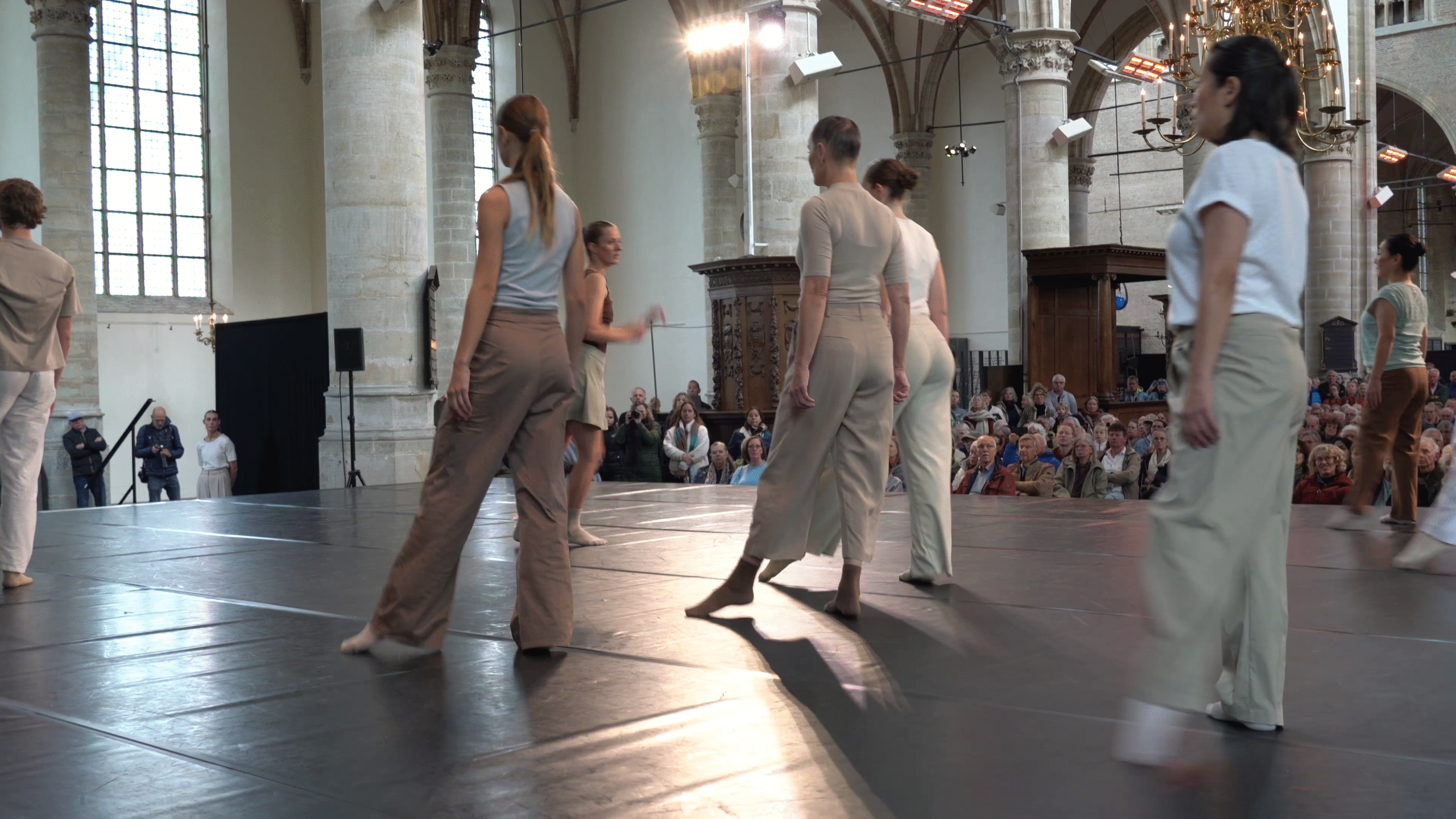 Verschillende dansers in neutrale kleding voeren een moderne dans uit op een podium in een historische kerk, omringd door een groot publiek dat aandachtig toekijkt.