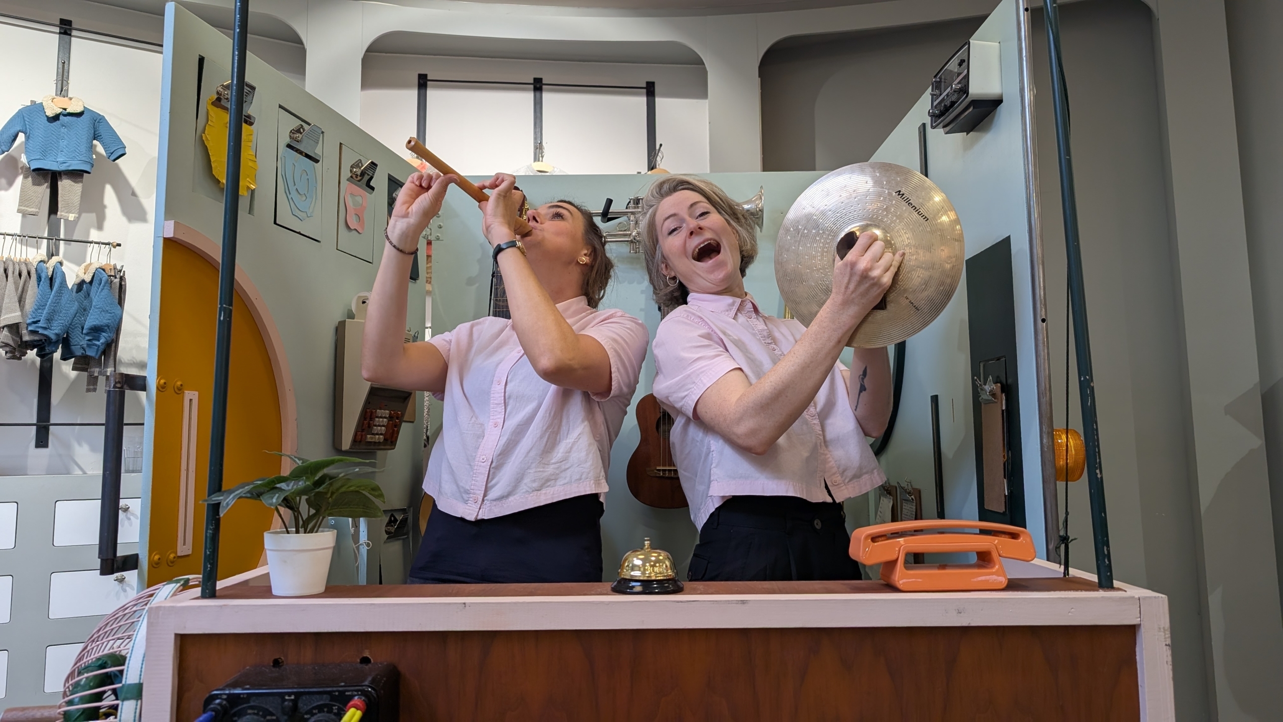 Twee vrouwen in roze shirts maken muziek; de een speelt een fluit en de ander slaat op een grote cimbaal, terwijl ze vrolijk poseren achter een toonbank met een bel en een oranje telefoon.