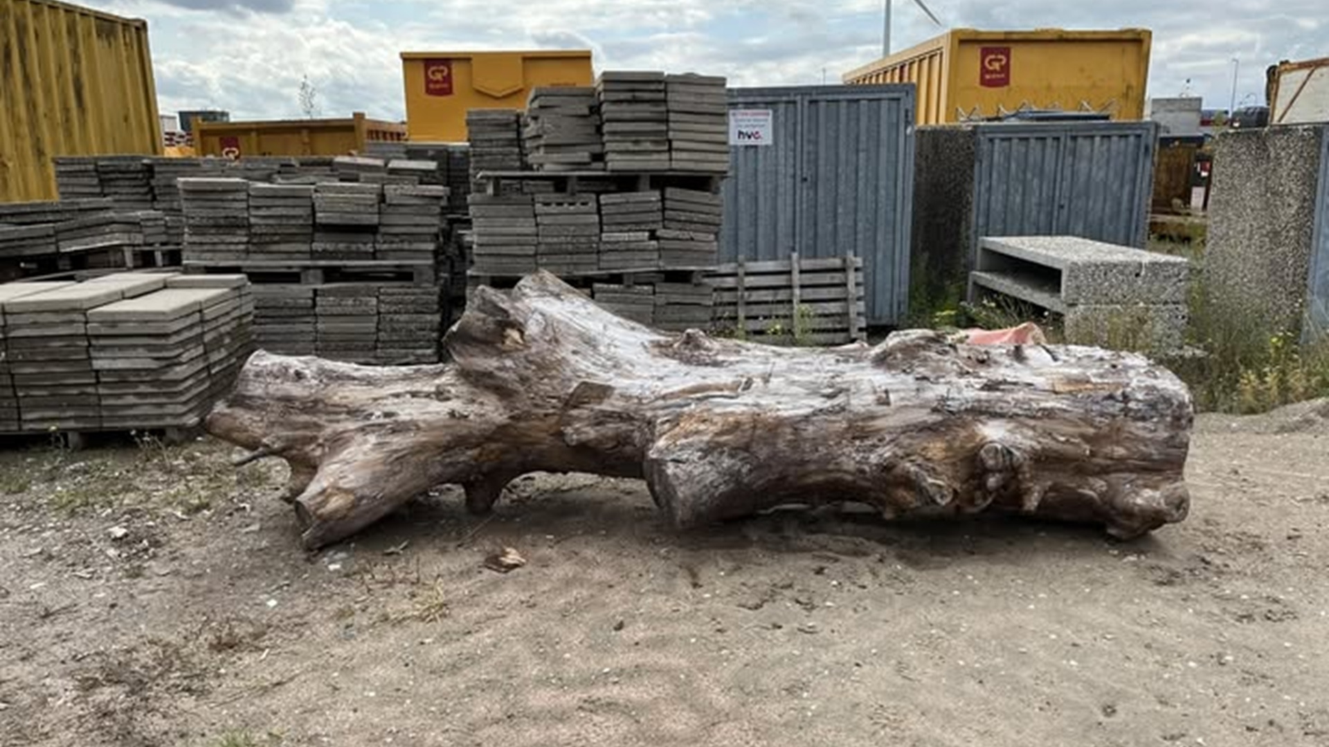 Grote houten boomstam ligt op een terrein met stapels stenen en ijzeren containers op de achtergrond.