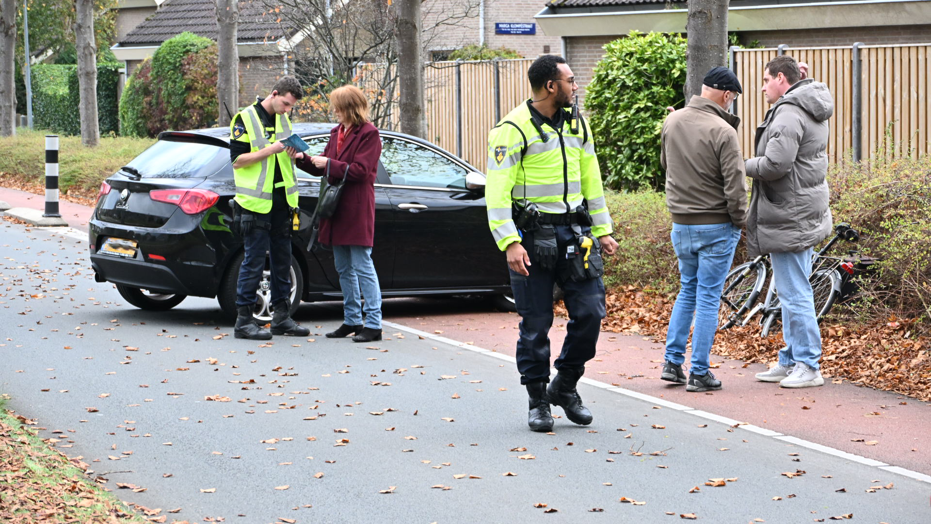 Twee agenten in gele veiligheidsvesten zijn bezig met mensen aan de kant van een weg. Een vrouw in een rode jas praat met een van de agenten naast een zwarte auto, terwijl twee mannen naast een fiets staan te praten met de andere agent. Er liggen bladeren op de grond, wat wijst op een herfstsfeer.