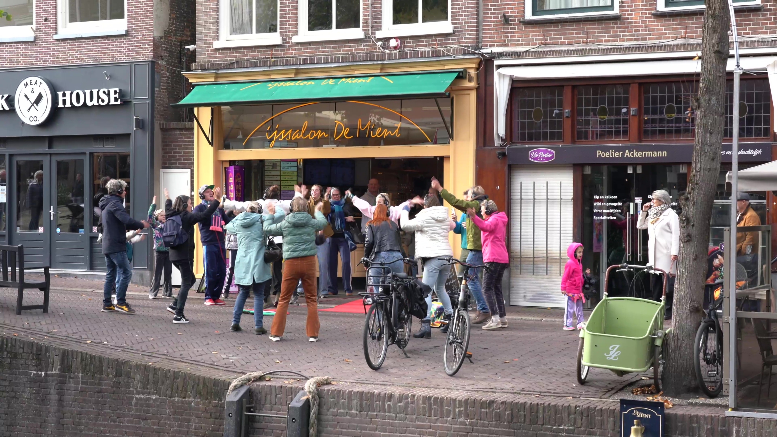 Een groep mensen danst enthousiast voor een ijssalon met een groene luifel, langs een straat met fietsen geparkeerd op de voorgrond.