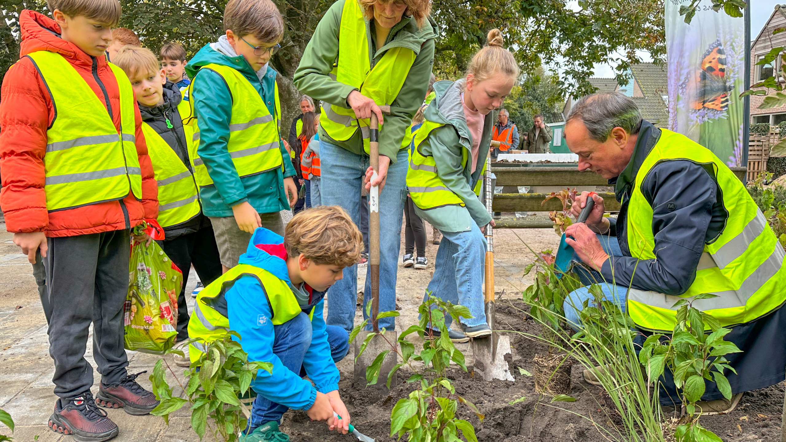 Kinderen en volwassenen dragen gele veiligheidshesjes terwijl ze gezamenlijk planten in de grond zetten in een tuinomgeving.