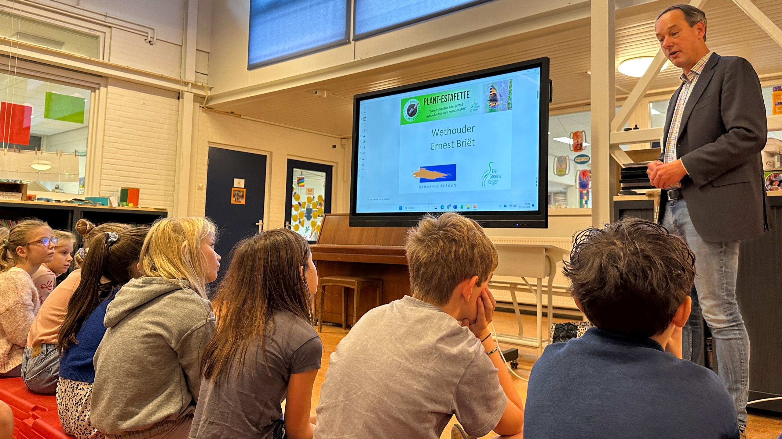 Groep kinderen luistert aandachtig naar een man die een presentatie geeft op een groot scherm in een klaslokaal.
