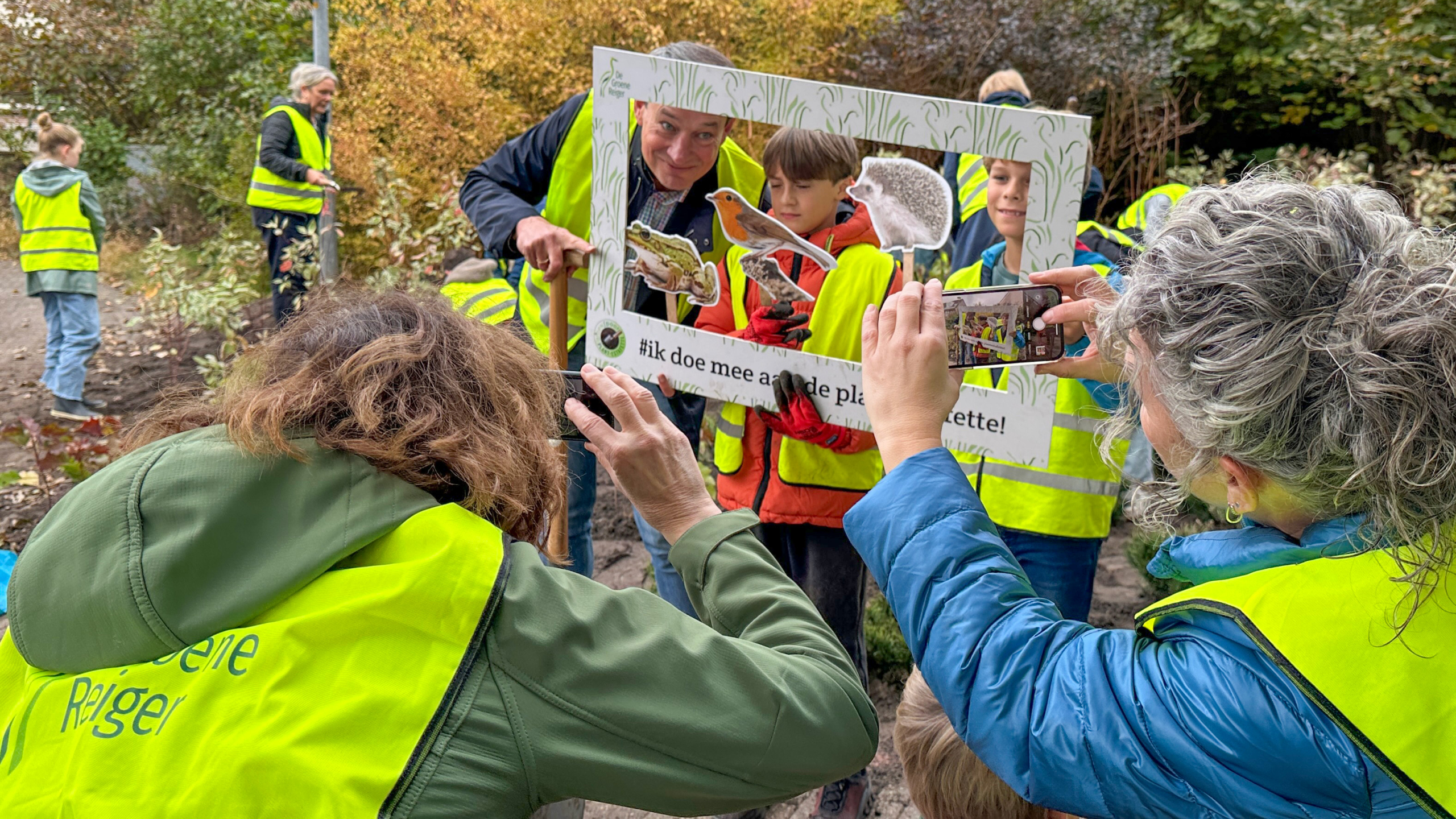 Mensen in gele veiligheidshesjes poseren buiten met een fotolijst waarop afbeeldingen van dieren zijn geplakt, terwijl iemand een foto maakt met een telefoon.