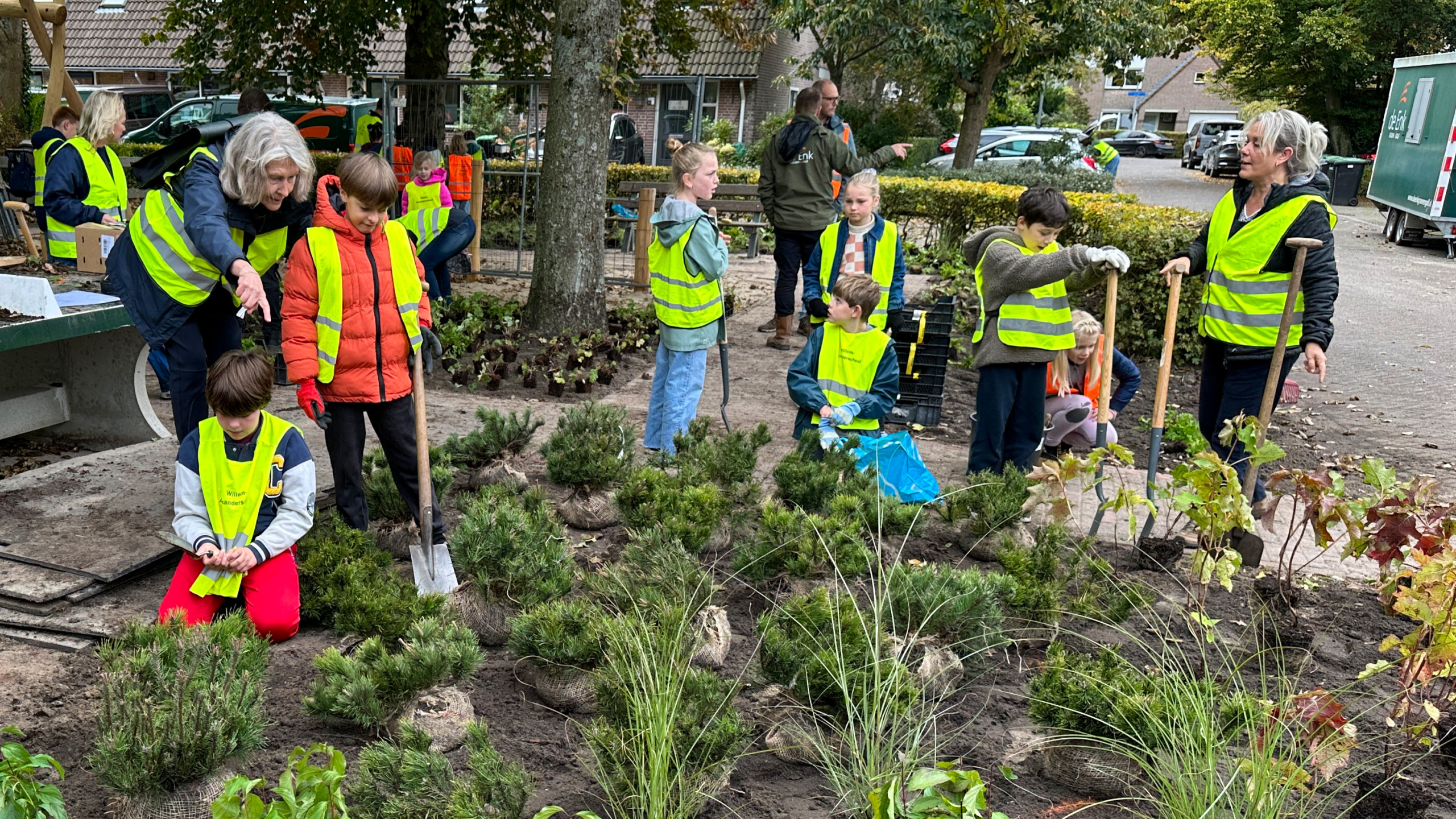Mensen, voornamelijk kinderen, dragen gele veiligheidsvesten en planten groene struiken in een tuinomgeving.