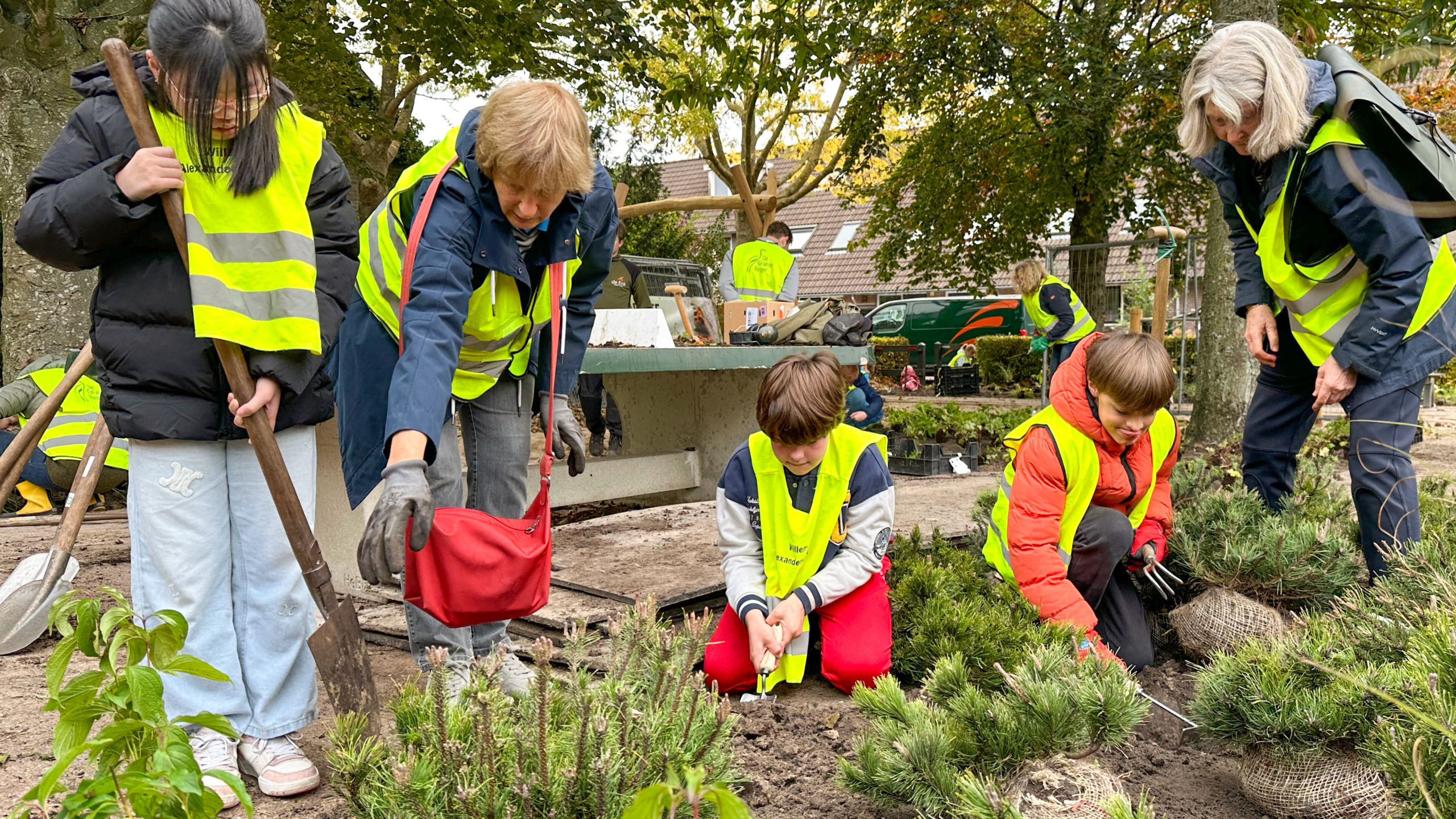 Mensen in gele veiligheidsvesten planten struiken in een tuin, geholpen door volwassenen in een buitenomgeving met bomen op de achtergrond.