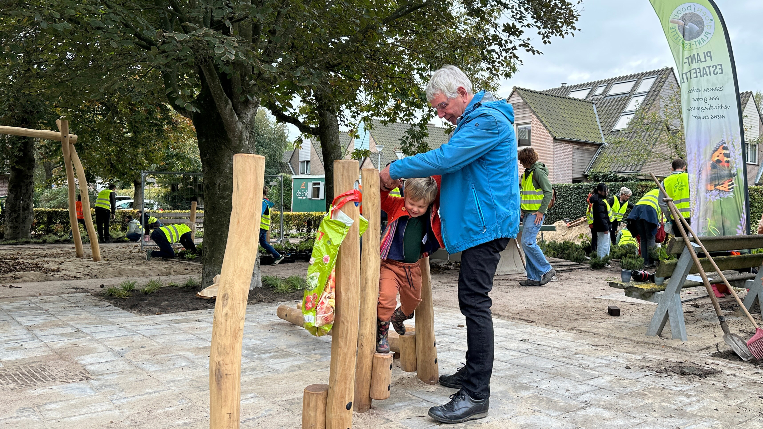 Een oudere man helpt een kind over een houten klimconstructie in een speeltuin, terwijl op de achtergrond mensen met gele veiligheidshesjes bezig zijn met tuinwerkzaamheden.