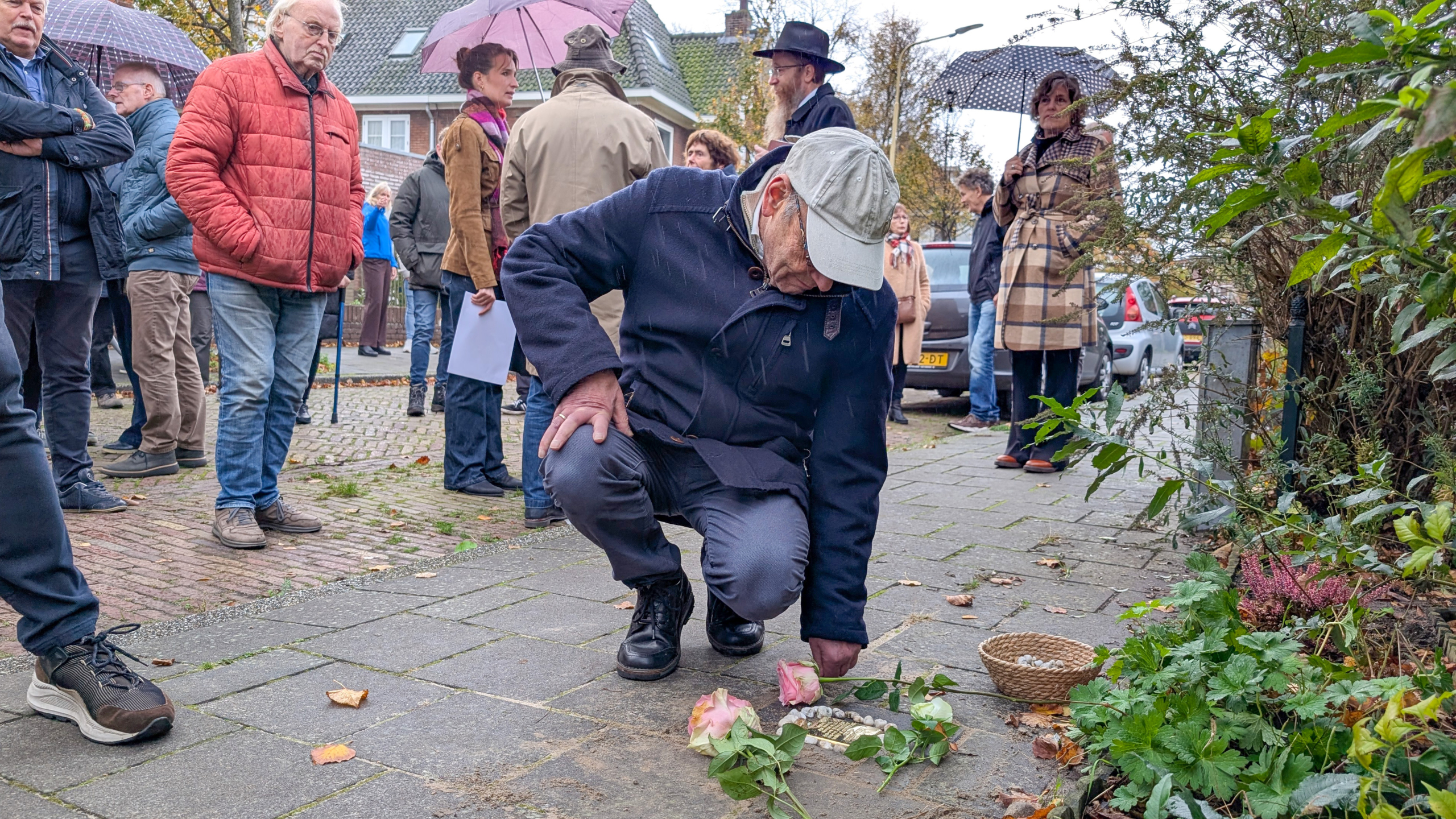 Een groep mensen staat op straat, waarvan een oudere man bukt om bloemen op de grond te leggen, omringd door anderen met paraplu's op een bewolkte dag.