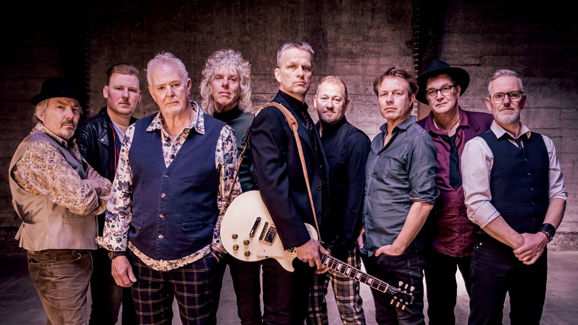 Een groep van negen mannen poseert, waarvan één een gitaar vasthoudt, in een ruimte met een industriële uitstraling.