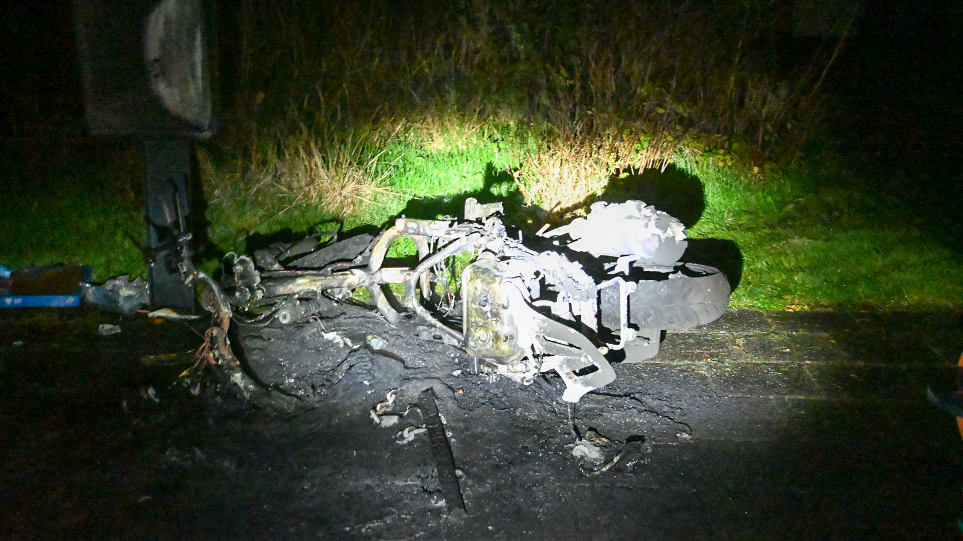 Verbrand wrak van een scooter op een donkere weg, omringd door gras, verlicht door een schijnwerper.