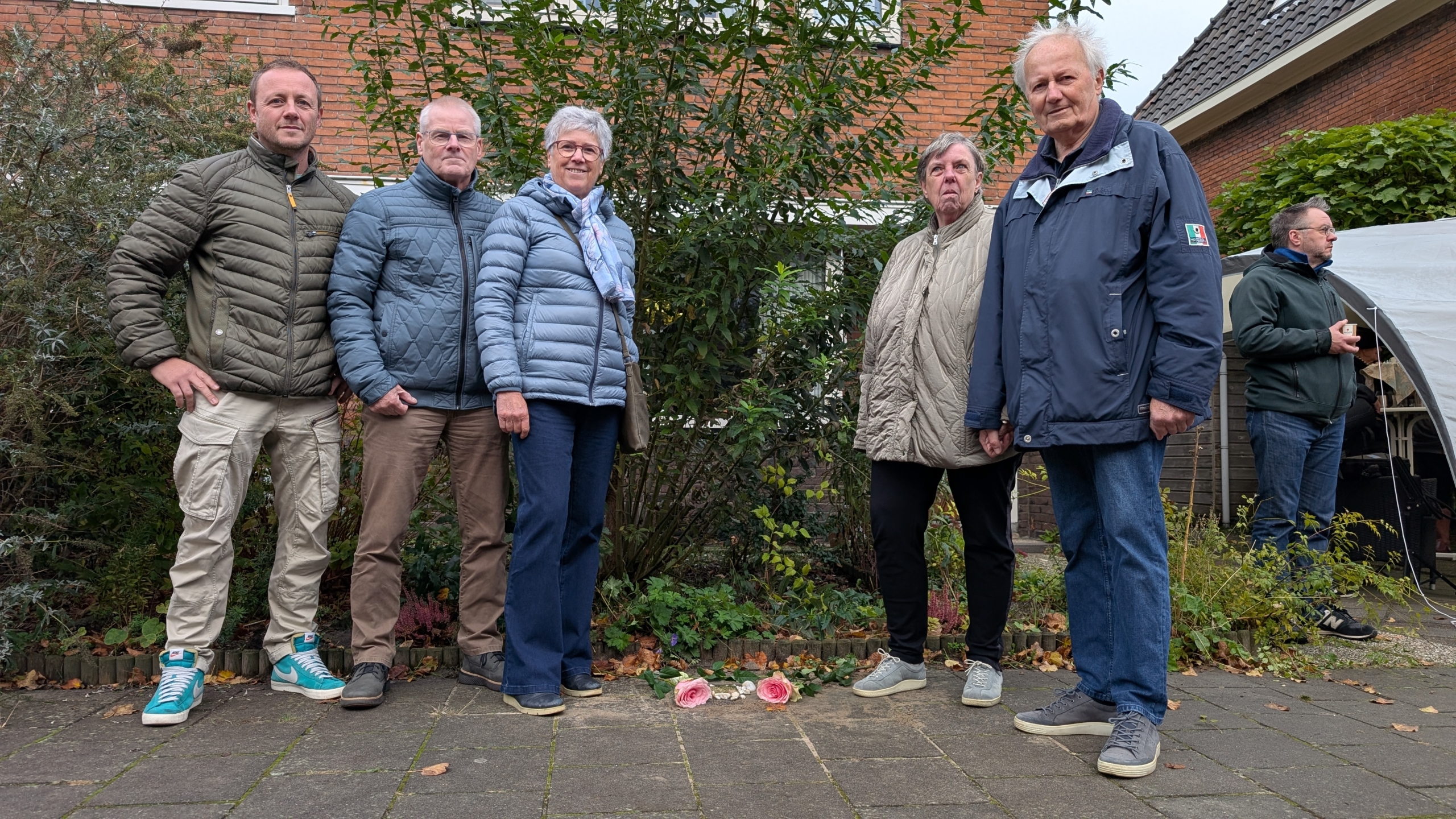 Groep van vijf mensen die buiten staan op een betegeld pad voor een huis, met bloemen op de grond in het midden.