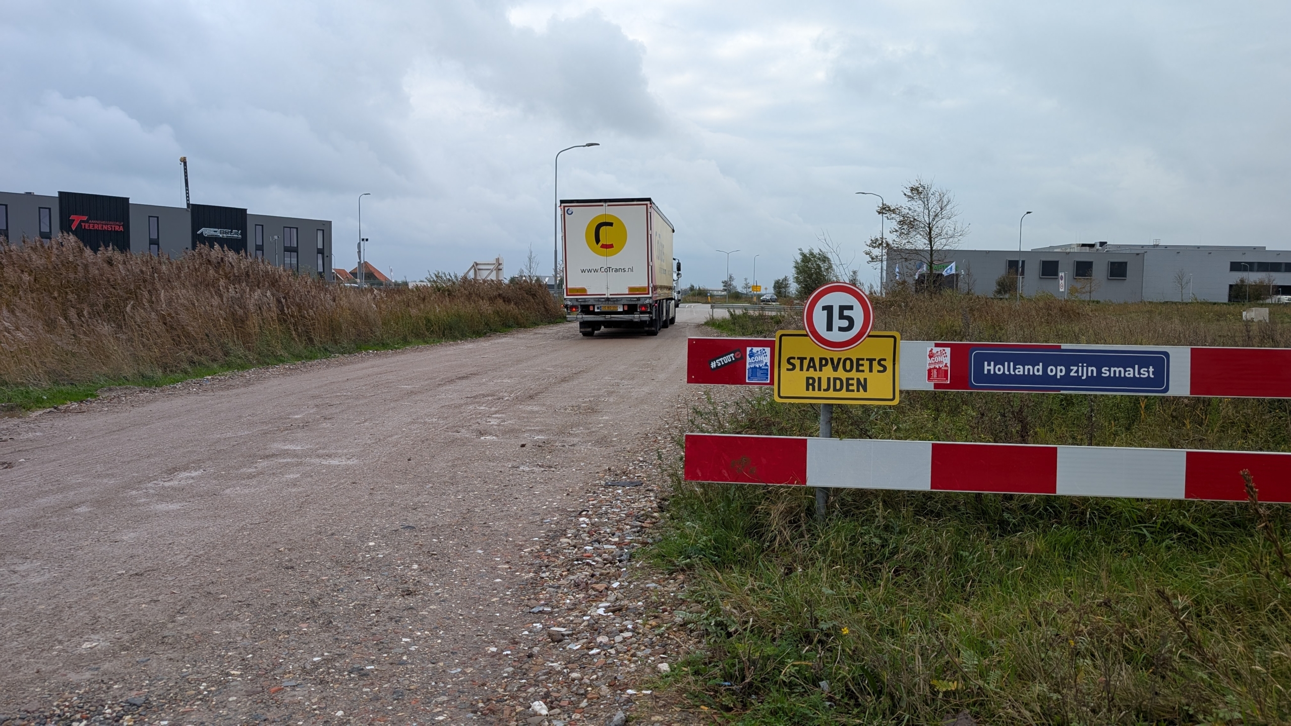 Een vrachtwagen rijdt over een onverharde weg naast hoge grassen, met links moderne gebouwen in de verte; op de voorgrond staat een bord met snelheidslimiet van 15 km/u en tekst "Stapvoets rijden" en "Holland op zijn smalst."