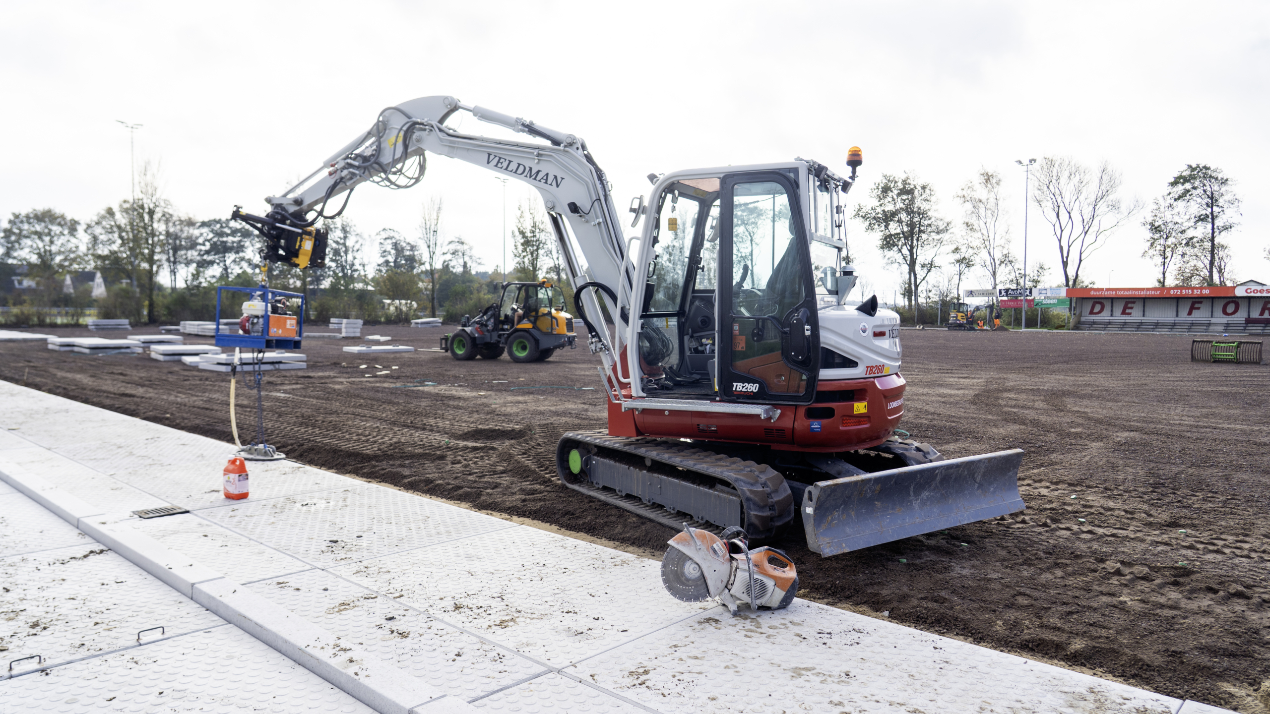 Graafmachine TB260 op een bouwterrein met een grijper aan de arm; een betonnen ondergrond en een lege tribune zijn zichtbaar.