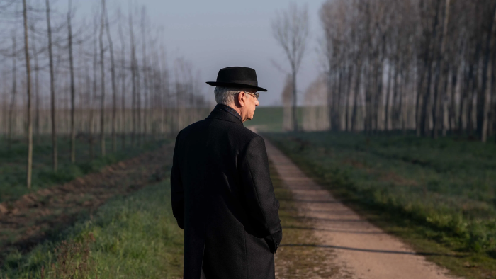 Man met hoed en overjas kijkt naar een landweg omgeven door bomen in een mistige ochtendomgeving.