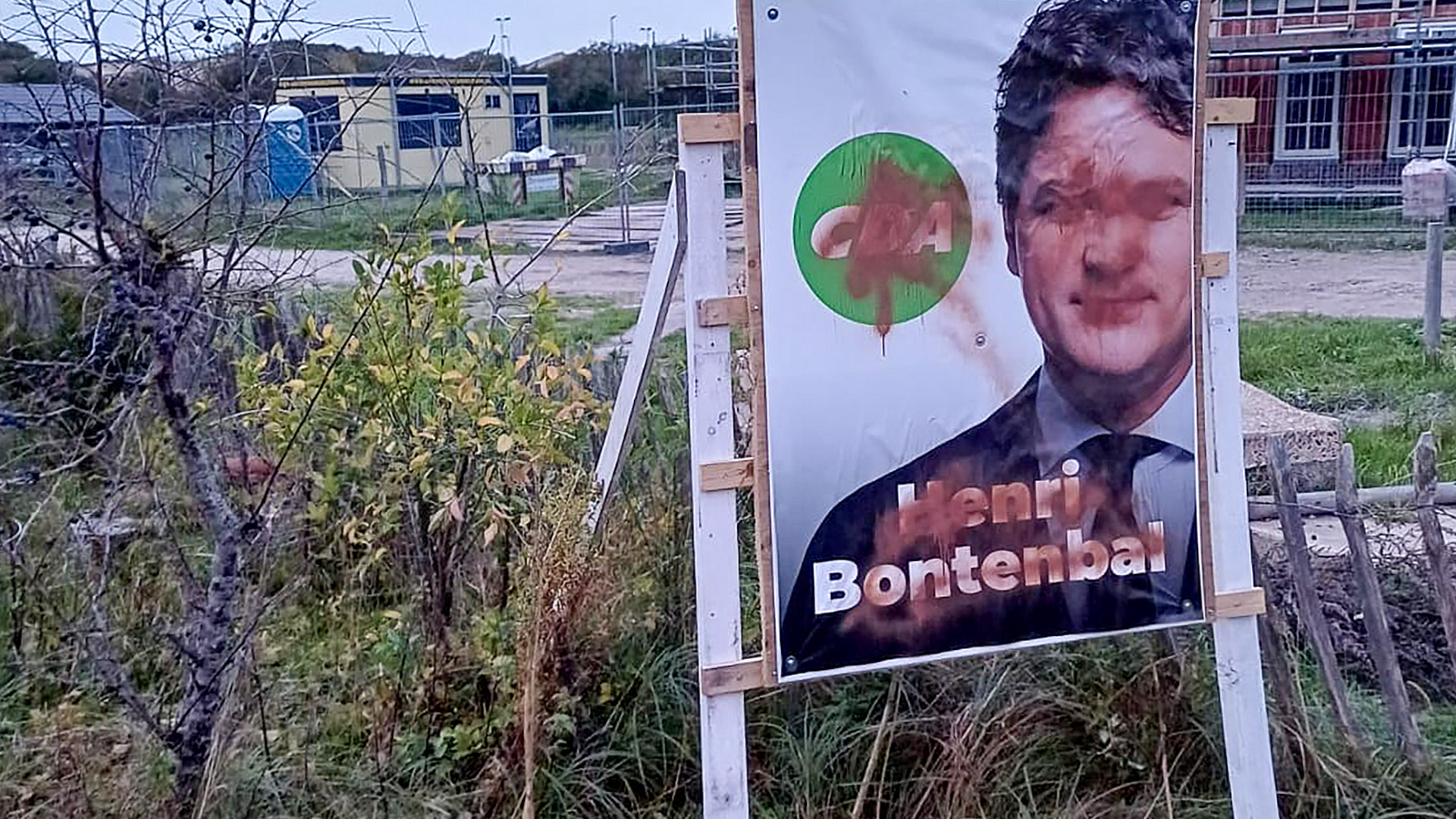 Verkiezingsposter van het CDA met een afbeelding van een man in pak bedekt met verf, geplaatst in een grasrijk gebied met een gebouw op de achtergrond.