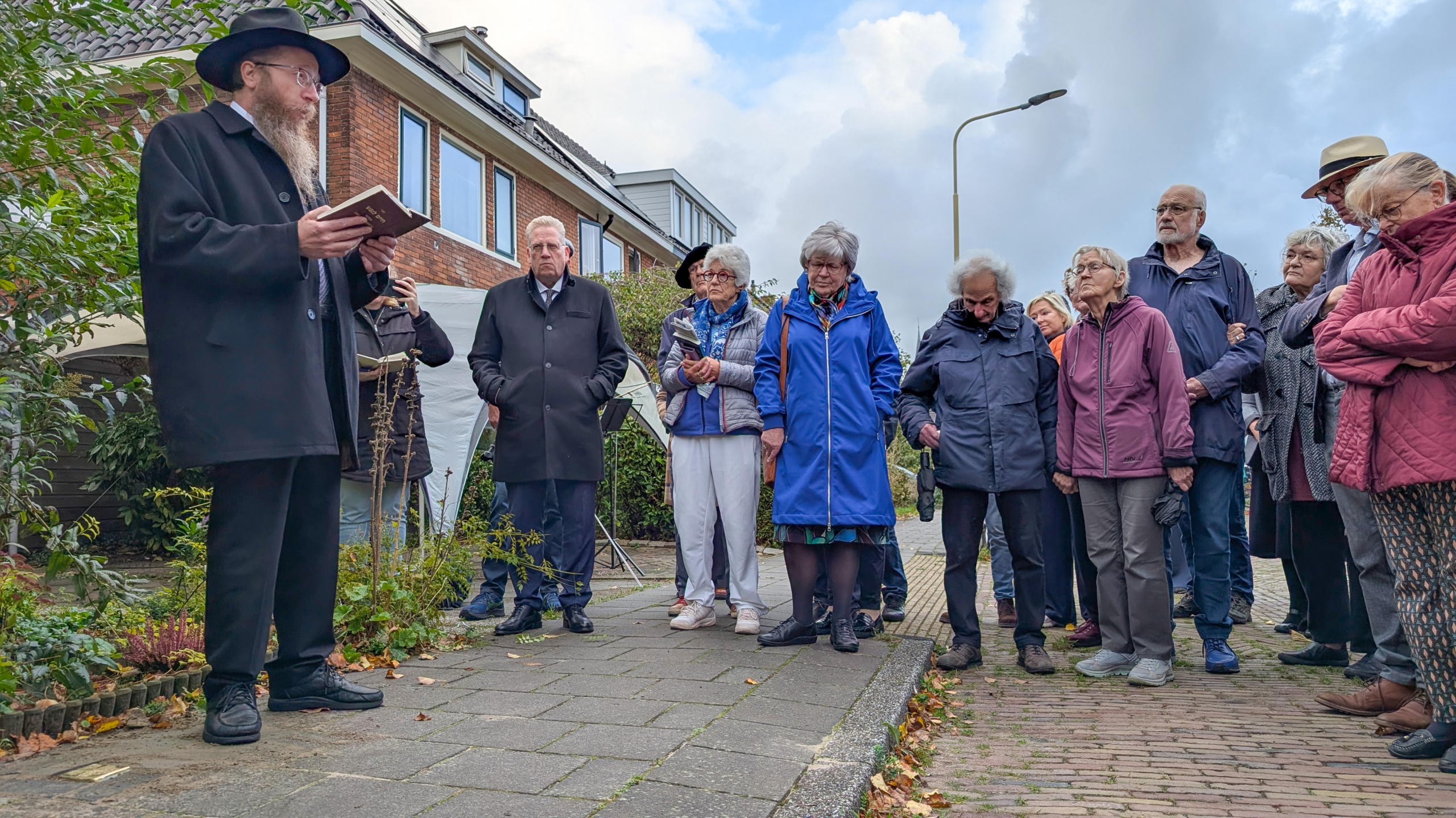 Een groep mensen luistert naar een man in traditioneel religieuze kleding die uit een boek voorleest op een trottoir in een woonwijk.