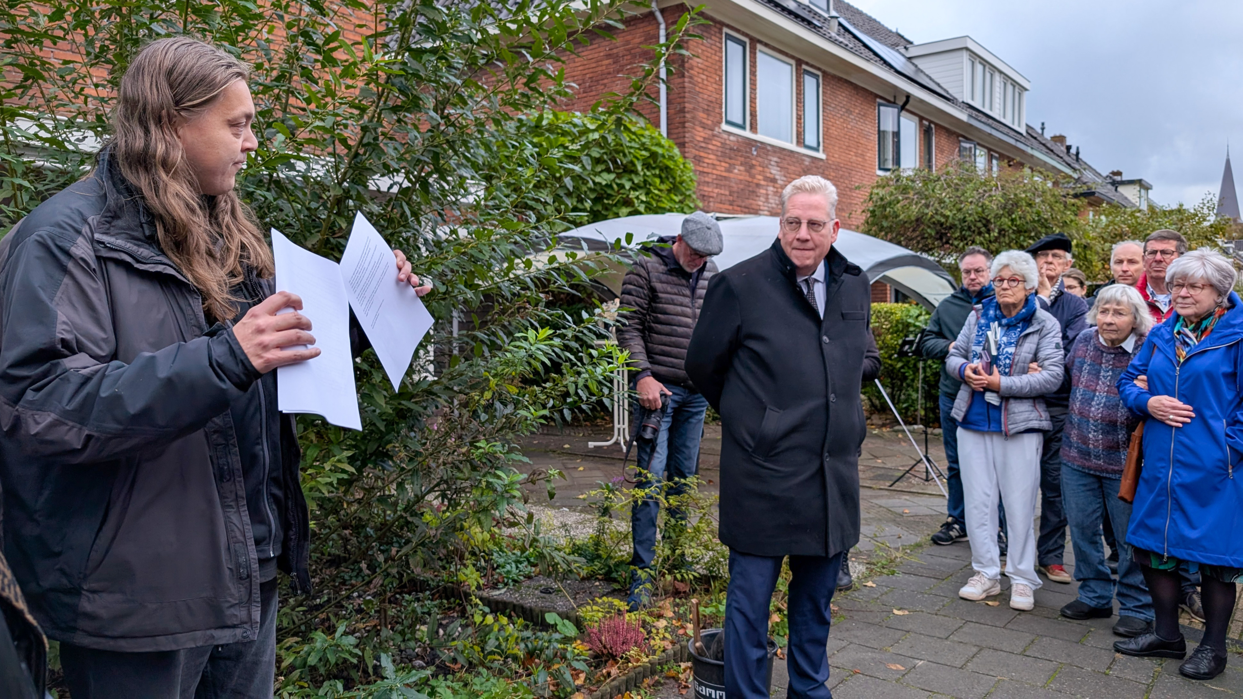 Een man met lang haar houdt enkele papieren vast terwijl hij buiten spreekt tot een groepje mensen die aandachtig luisteren, naast een gebouw.