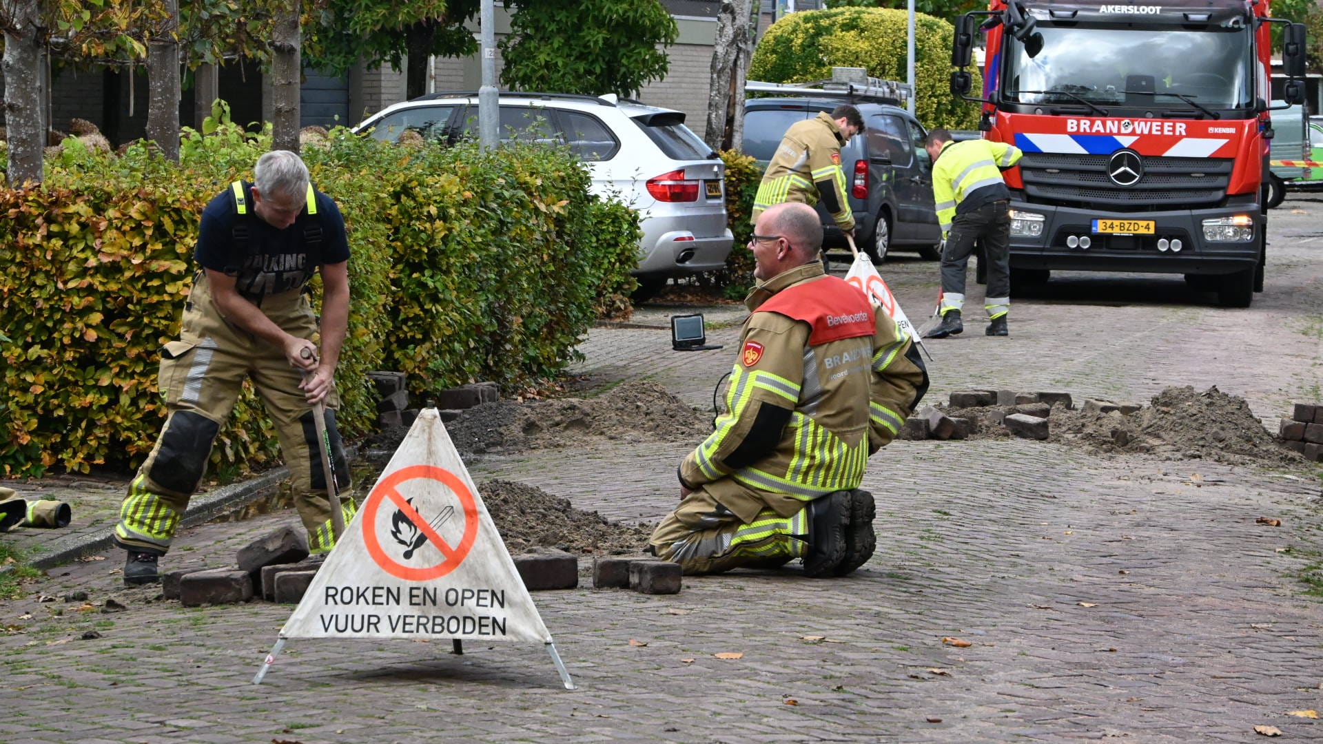 Brandweermannen werken aan een opengebroken weg, met een verbodsbord voor roken en open vuur. Op de achtergrond staat een brandweerwagen geparkeerd.