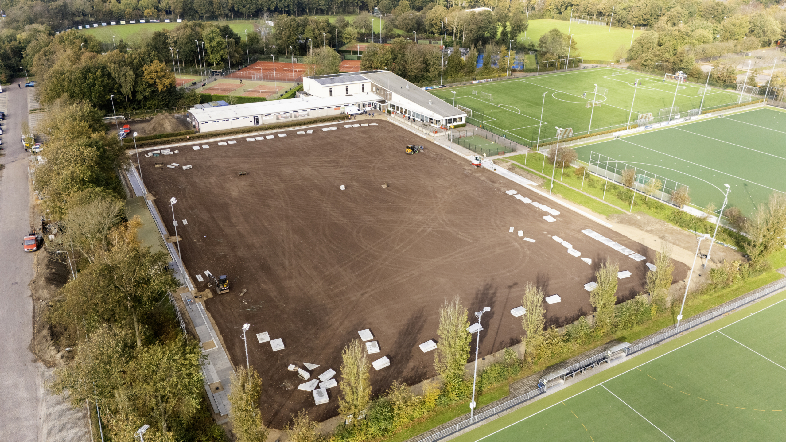 Luchtfoto van een sportcomplex met een groot bruinkleurig veld in aanbouw, omringd door bestaande groene grasvelden en tennisbanen. Er staat een gebouw naast de velden en enkele voertuigen en stenen platen zijn zichtbaar op het bruine veld.