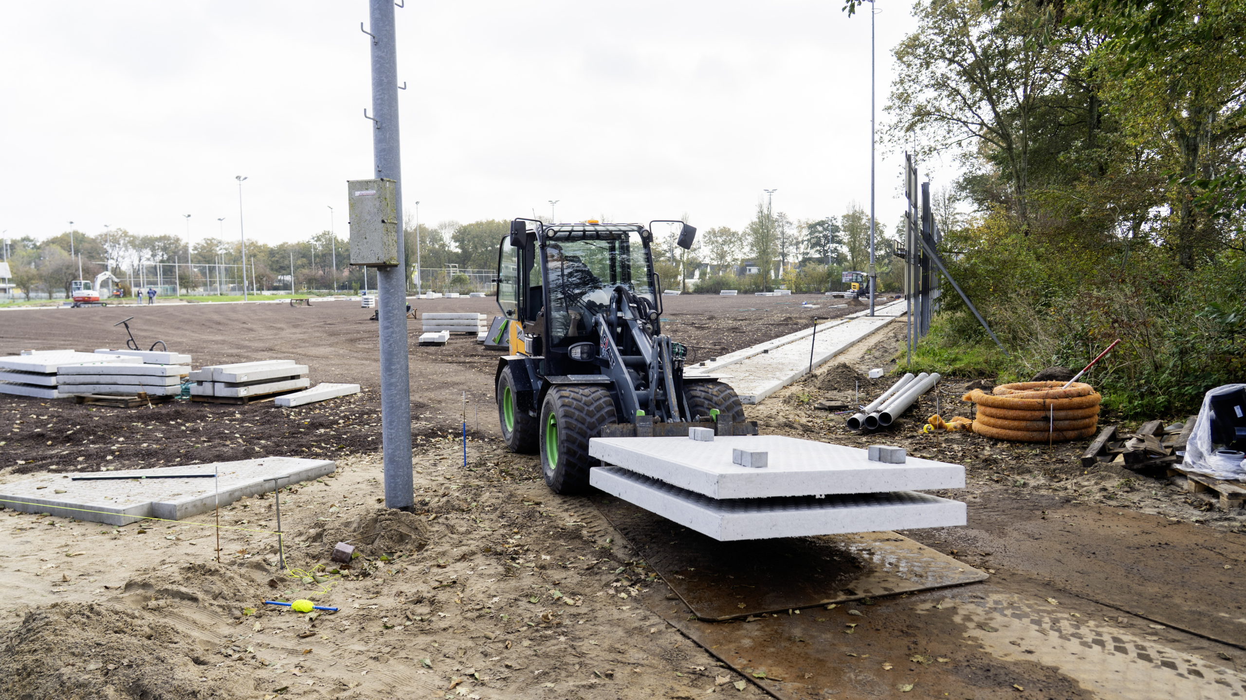 Een kleine wiellader vervoert betonnen platen op een bouwplaats met zandgrond en stapels platen op de achtergrond, omgeven door bomen en gereedschap zoals buizen en kabelrollen.