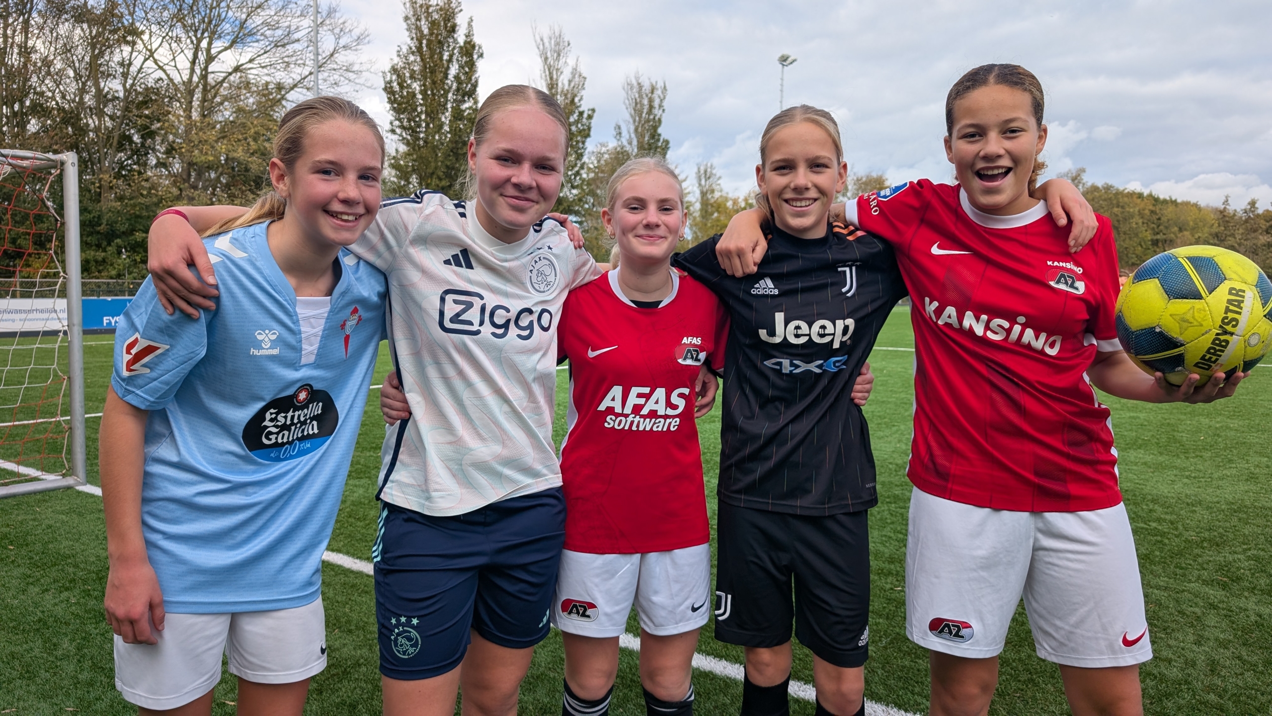 Vijf jonge voetballers in verschillende teamshirts poseren vrolijk op een voetbalveld, met één persoon die een bal vasthoudt.